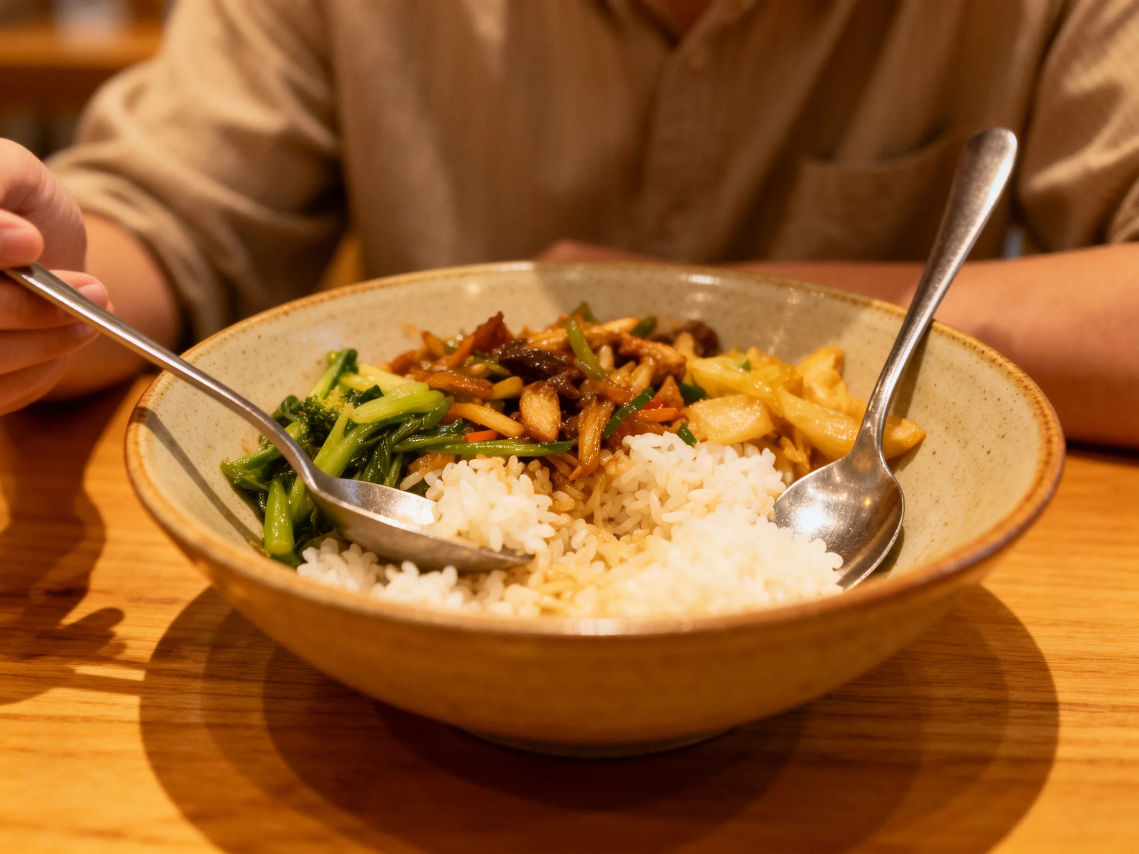 A bowl of rice, stir-fry, and broccoli with two metal spoons on a wooden table