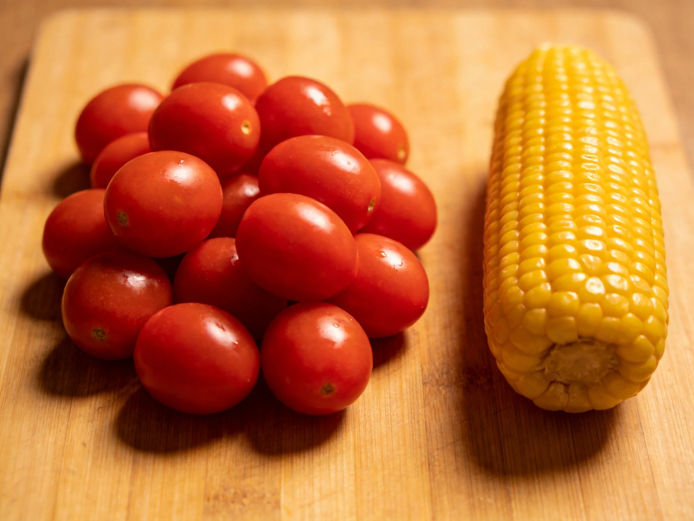 A cluster of red cherry tomatoes beside a single yellow corn ear on a wooden cutting board