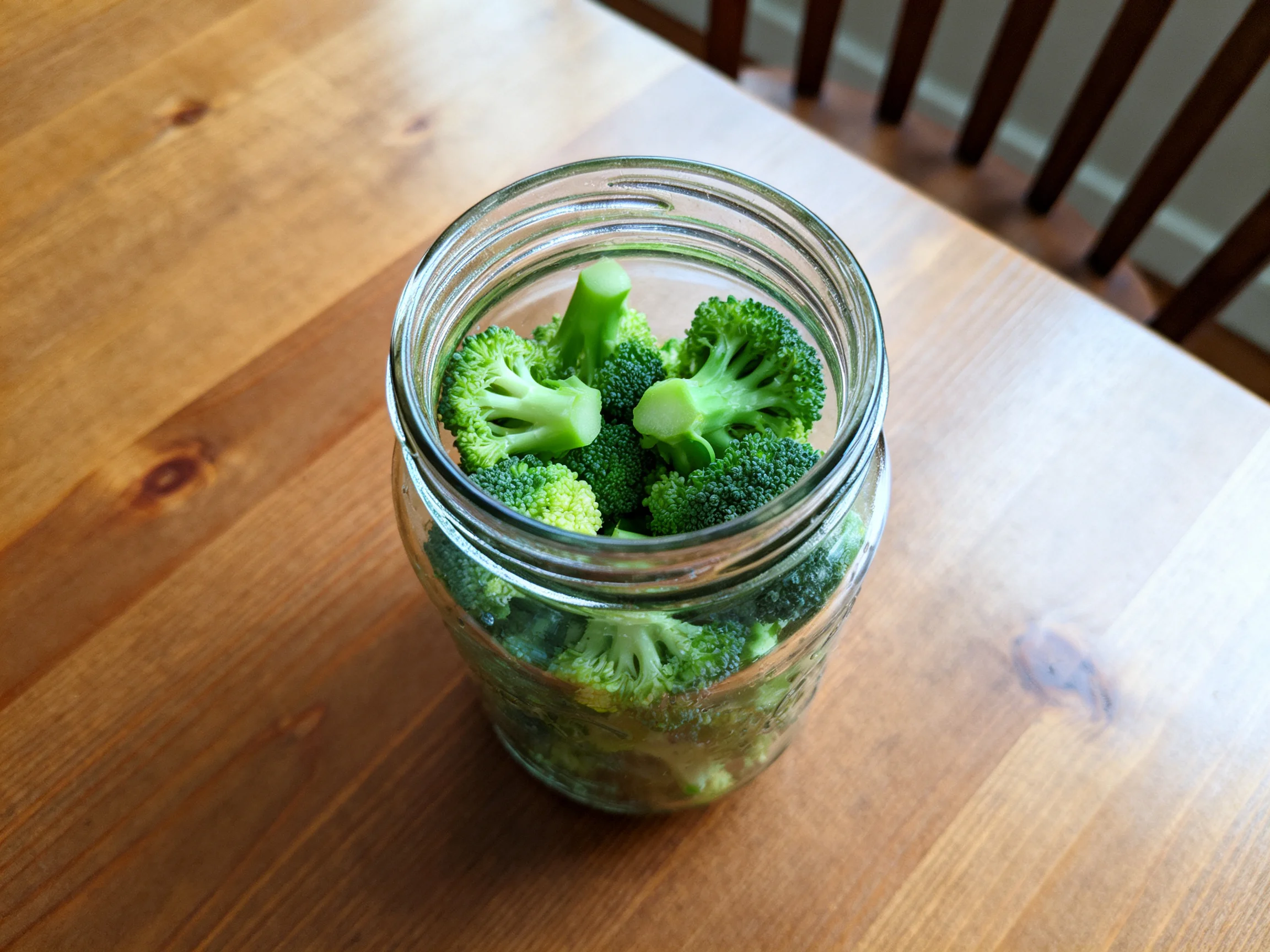 A glass jar filled with fresh green broccoli sits on a wooden table