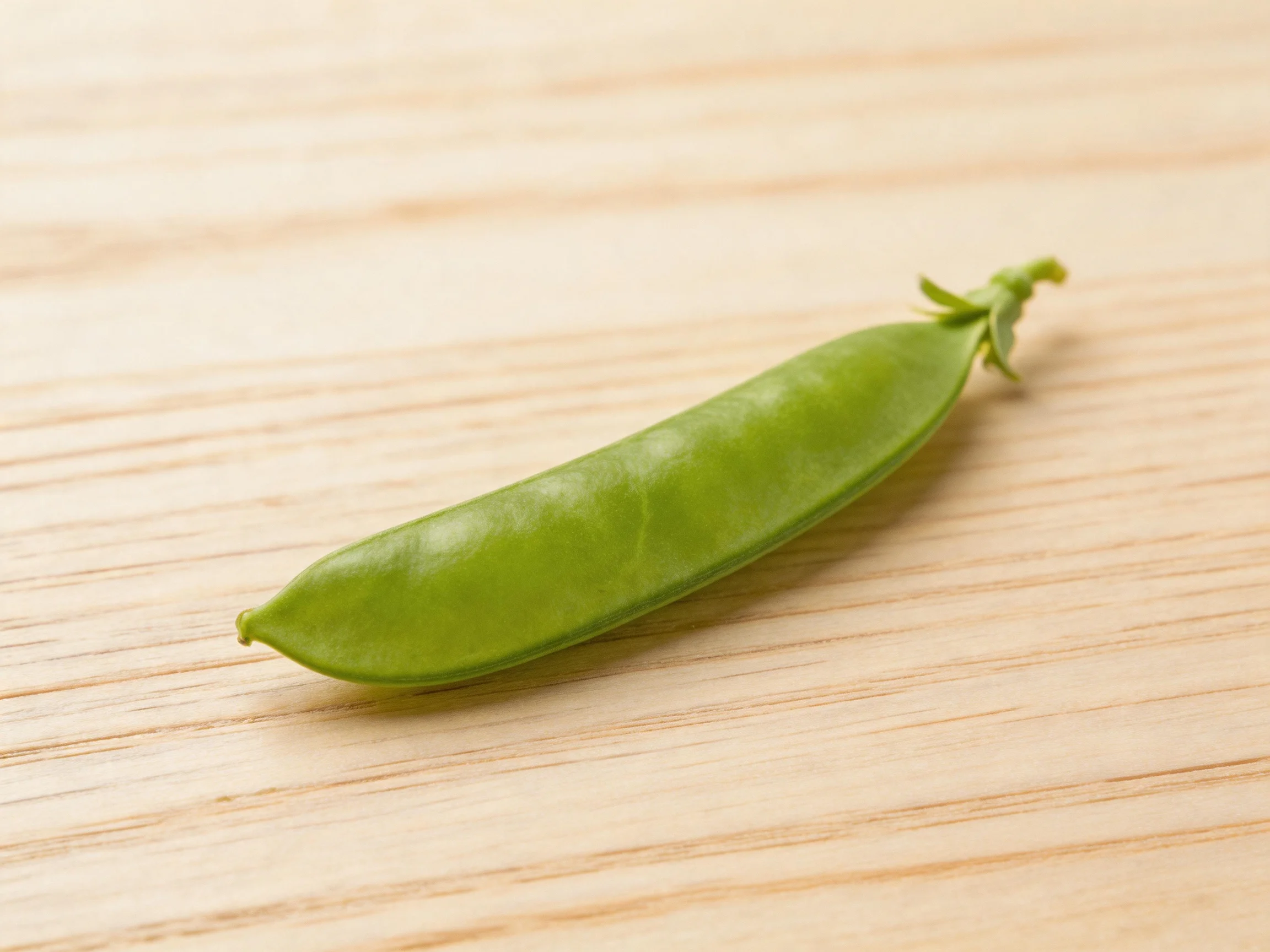 A single vibrant green pea pod rests on a light wooden surface