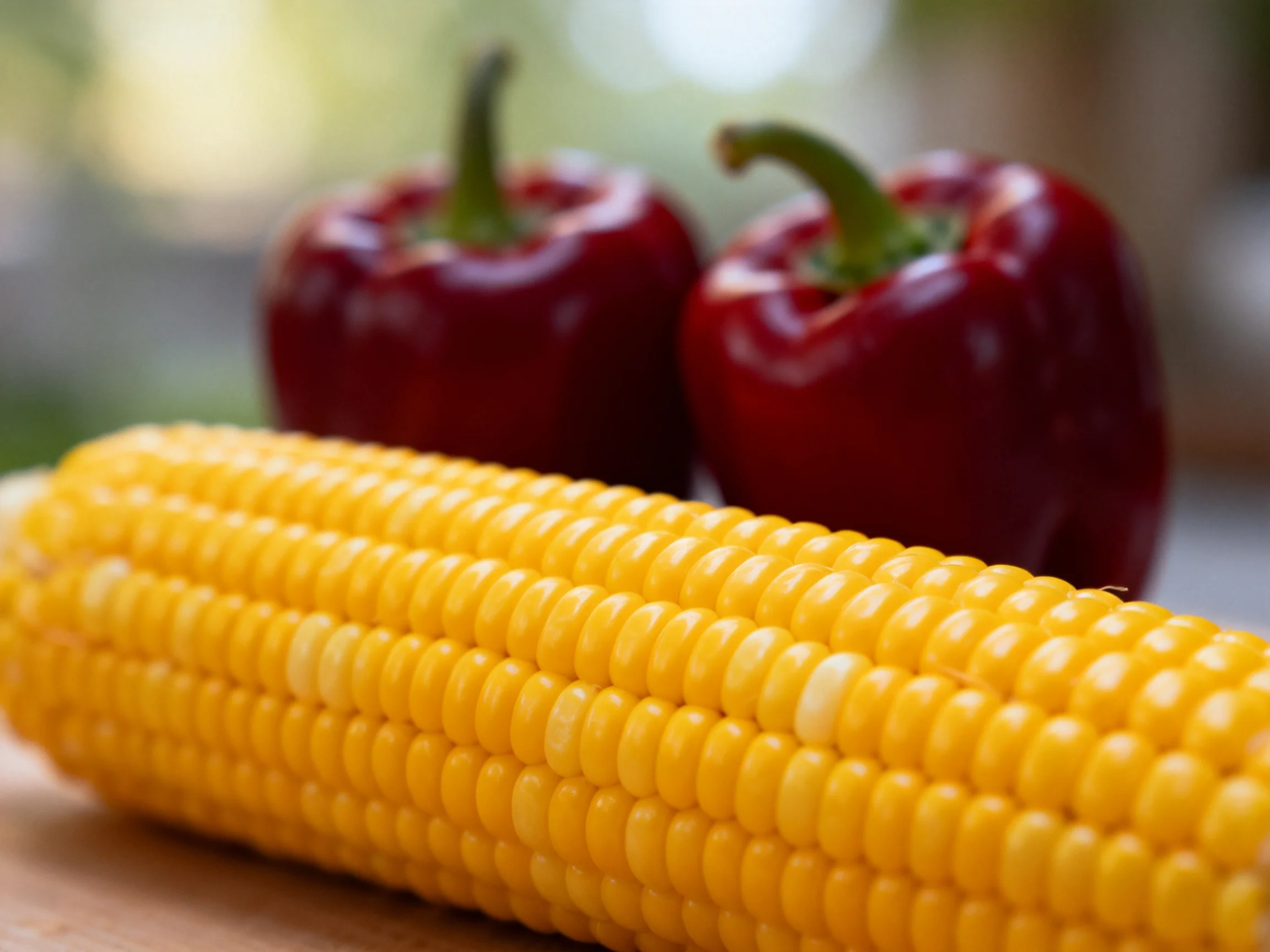 A vibrant yellow corn cob beside two glossy red bell peppers