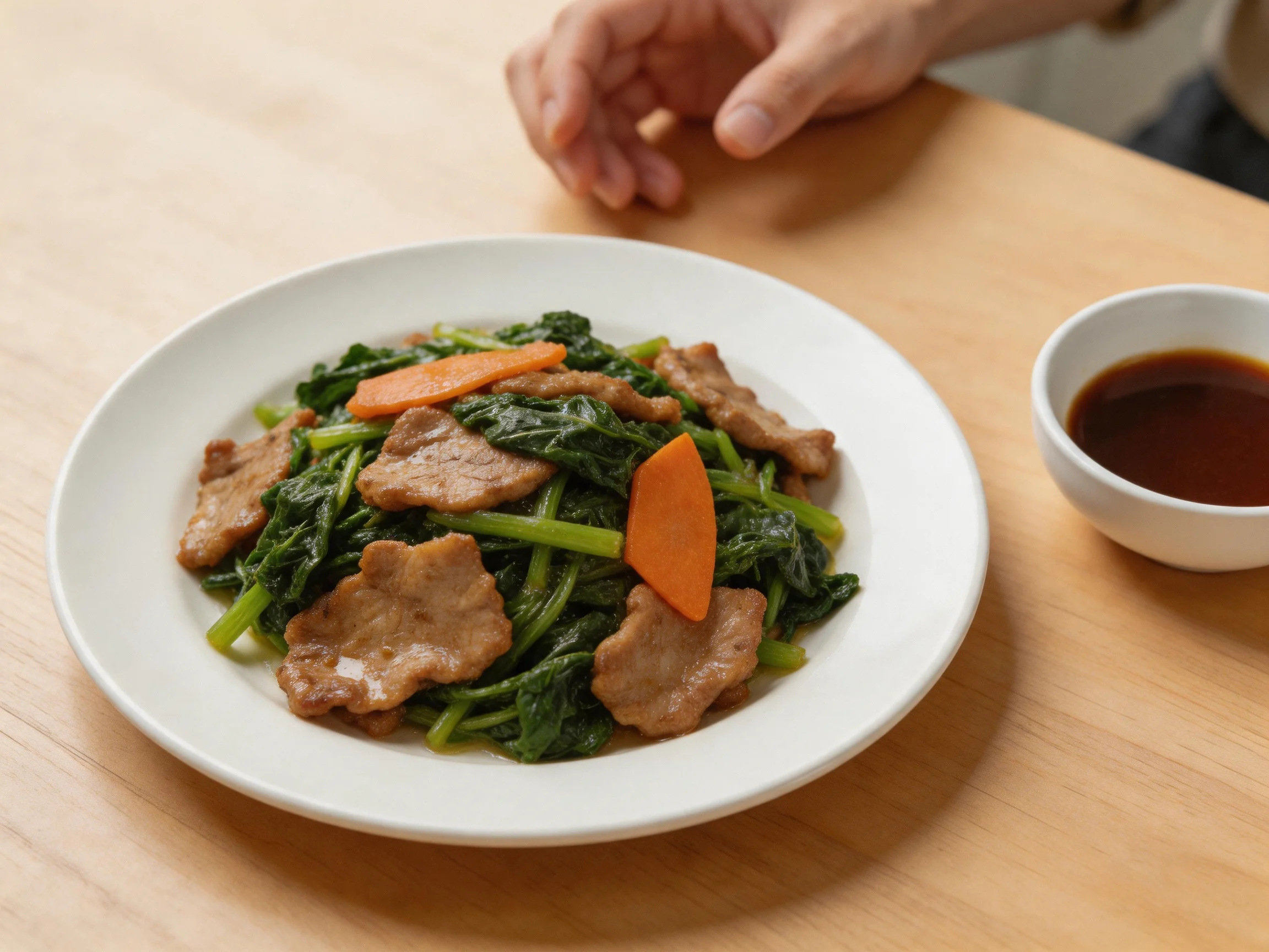 A white plate contains stir - fried pork, leafy greens, and carrot slices beside a sauce dish