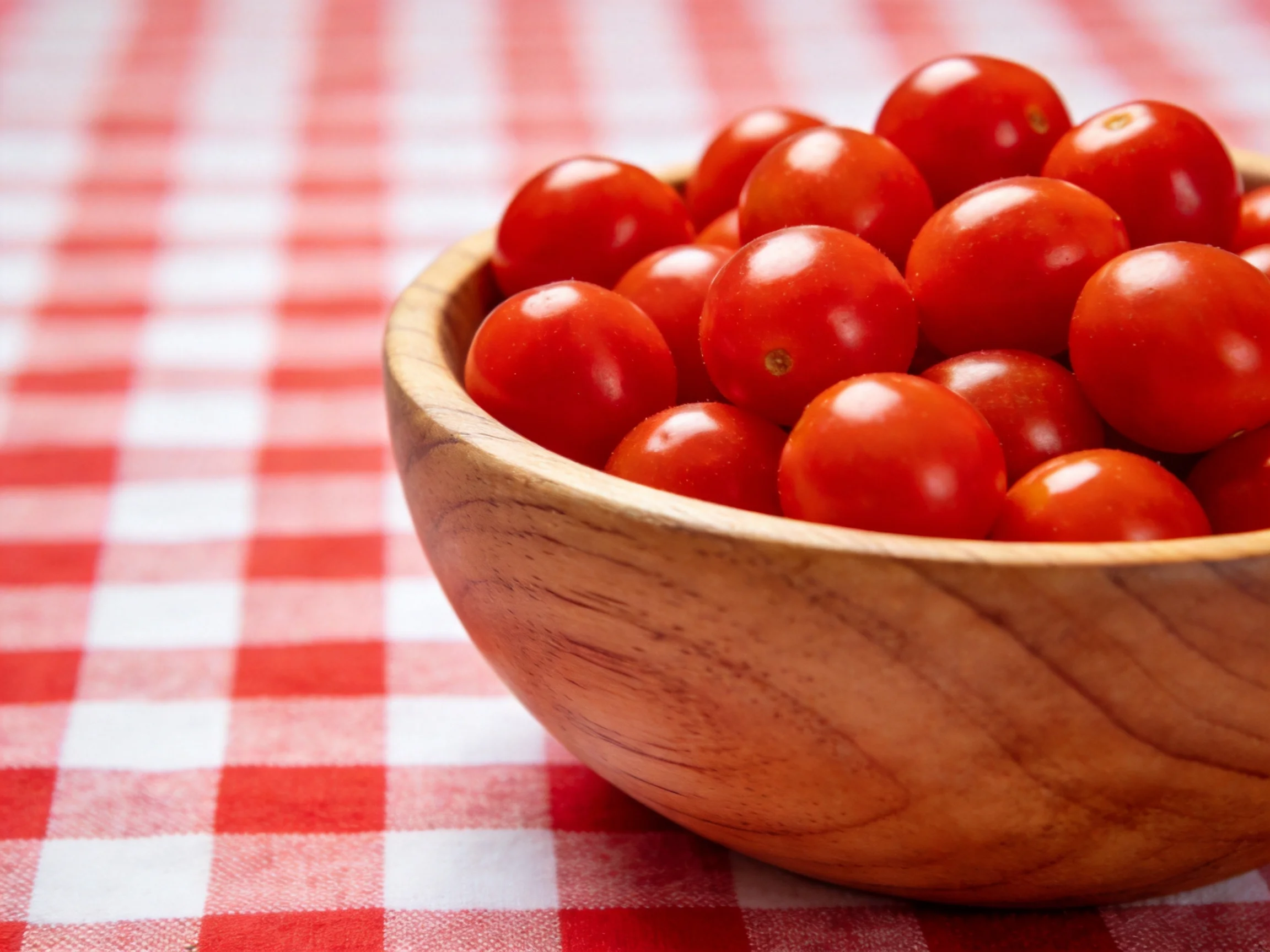 Cherry tomatoes overflow from a wooden bowl on a red-and-white checkered cloth