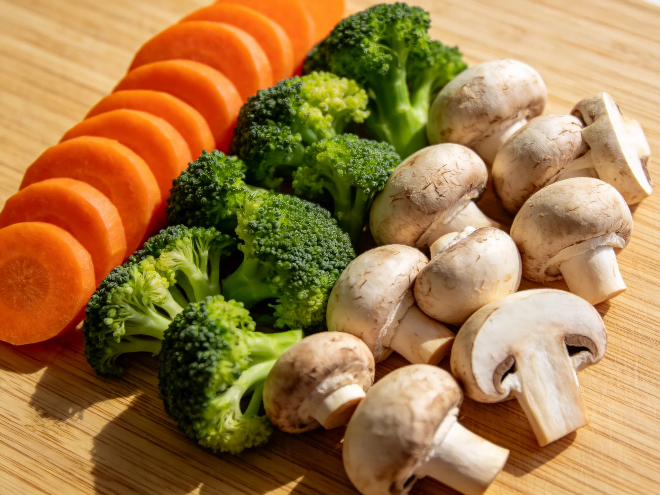 Fresh vegetable slices including carrot rounds, broccoli florets, and whole mushrooms on a wooden boa