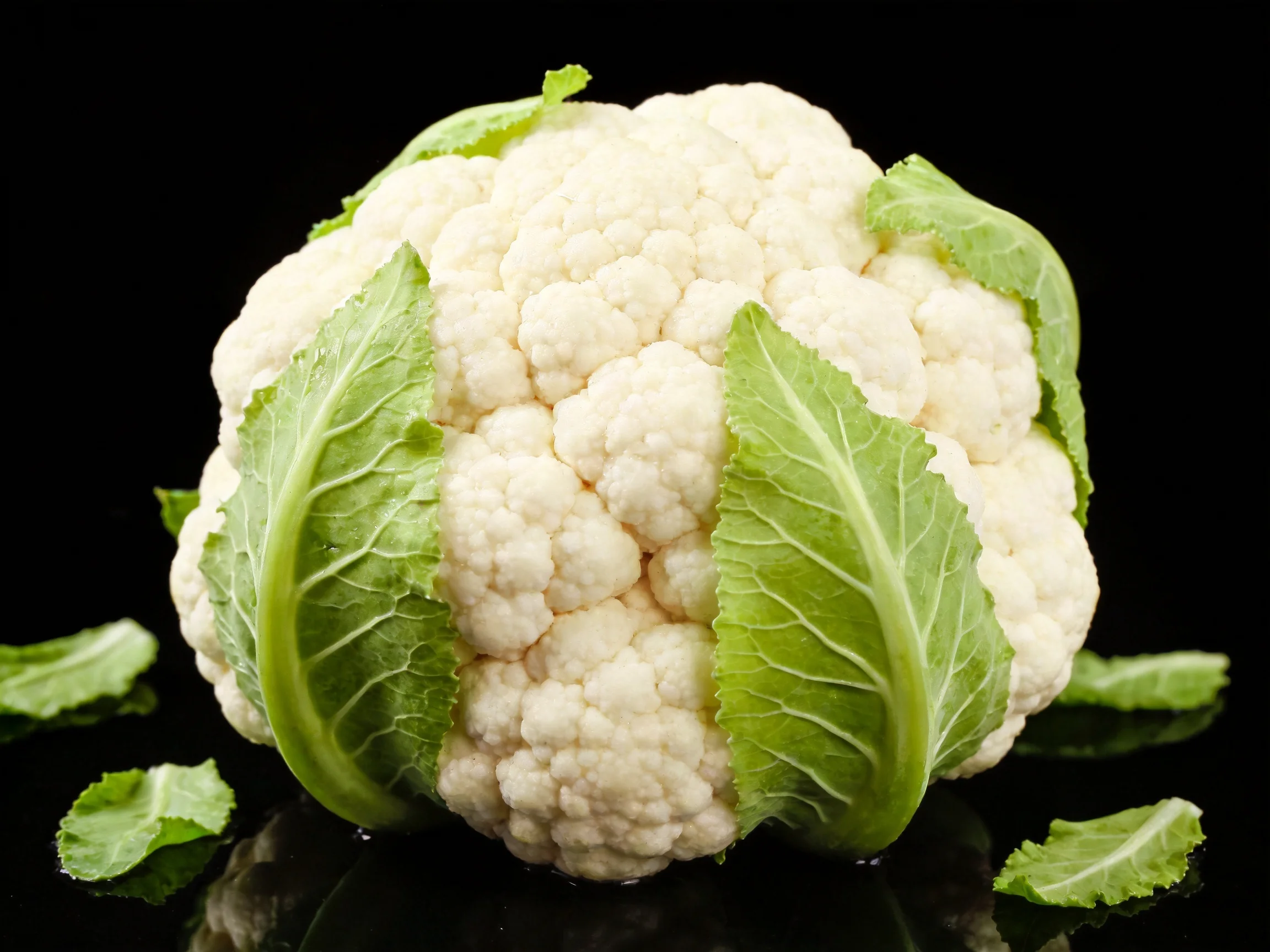 Fresh white cauliflower with vibrant green leaves on glossy black surface