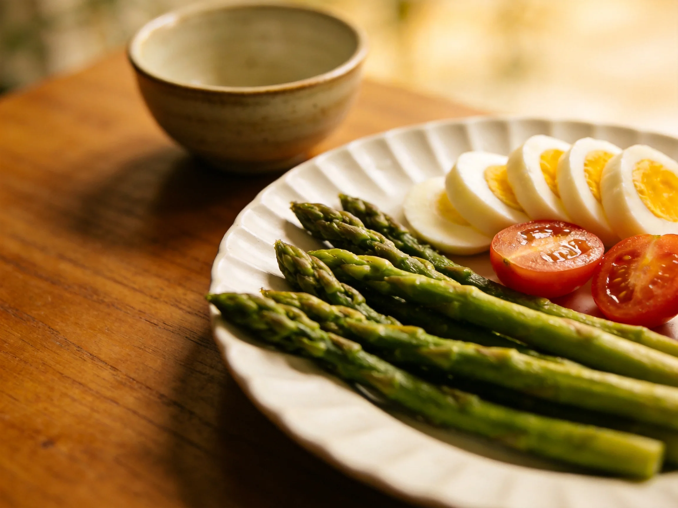 Grilled asparagus, sliced hard-boiled eggs, and cherry tomatoes on a plate beside a ceramic bowl