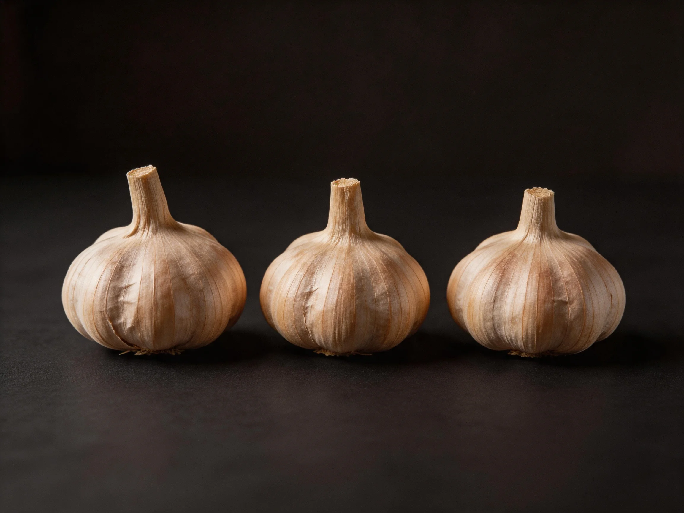 Three garlic bulbs with light brown papery skinson a dark surface