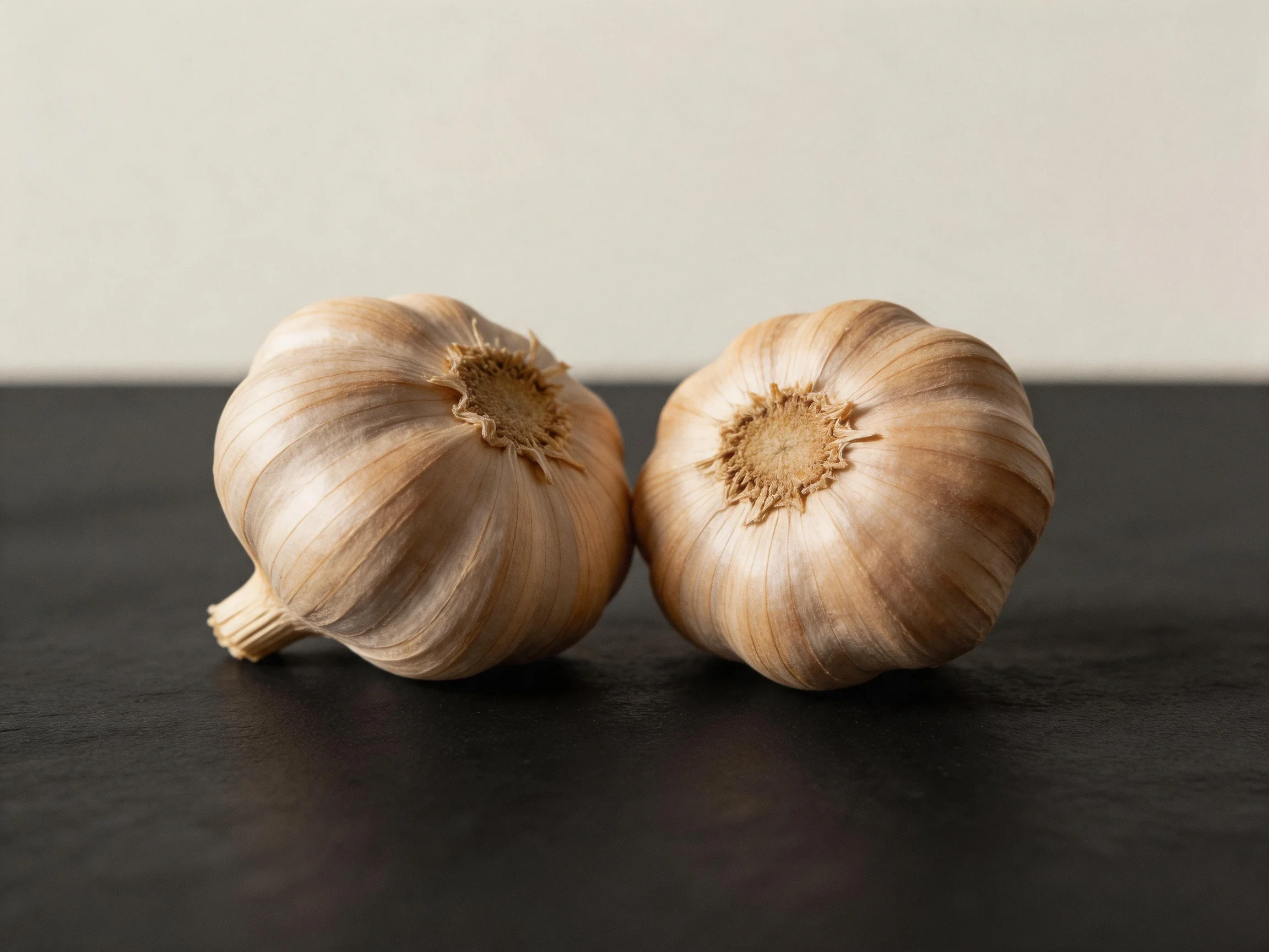 Two garlic bulbs with brown papery skins rest on a dark surface.