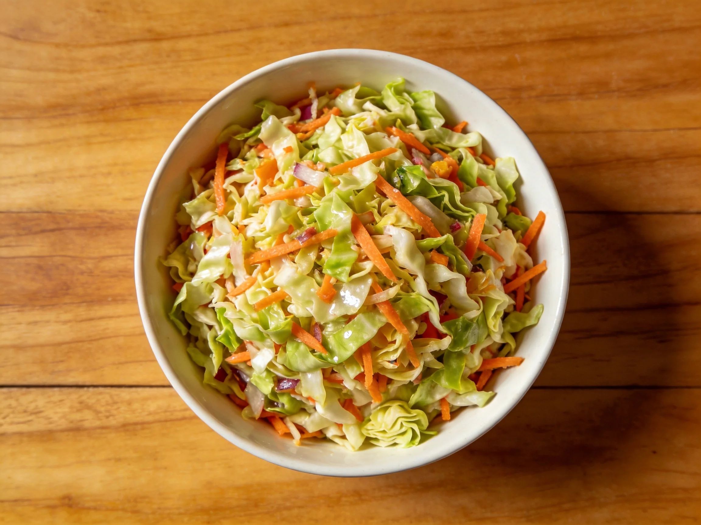 White bowl containing shredded cabbage and carrot slaw on wooden table