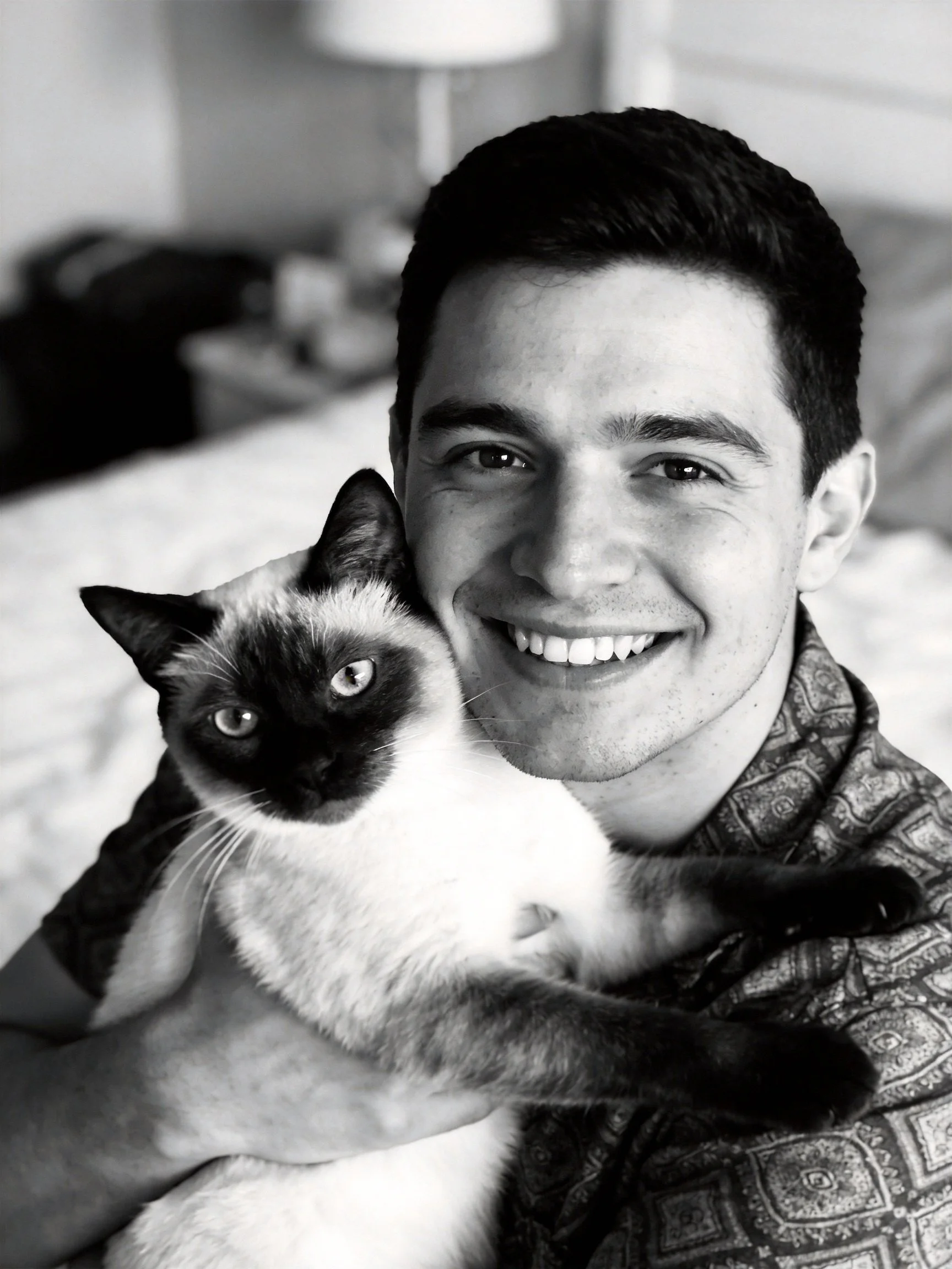 A man smiles while holding a black - and - white Siamese cat in a softly lit bedroom.