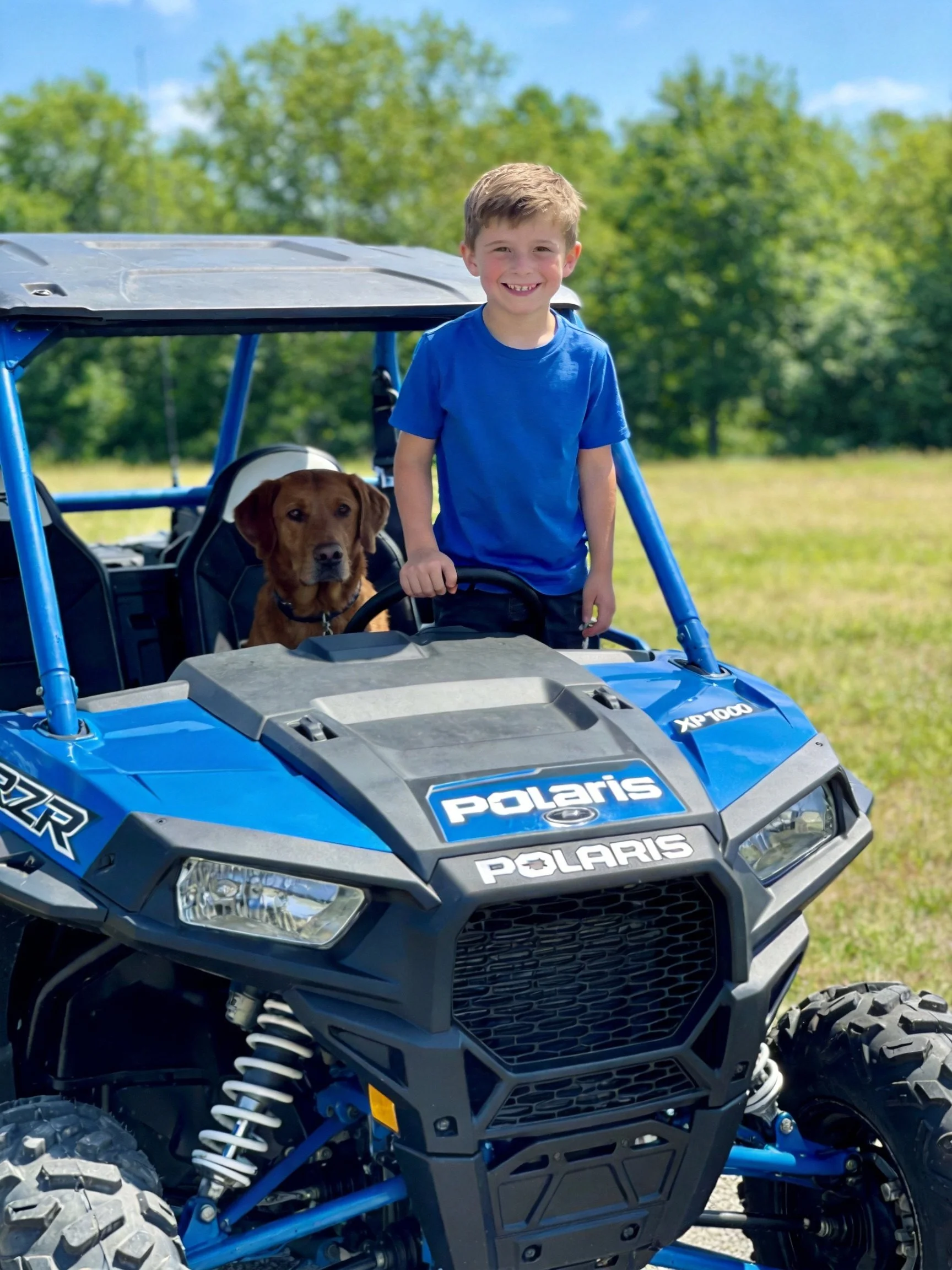 A boy and his dog are in a blue Polaris off-road vehicle in a grassy, tree-filled area.
