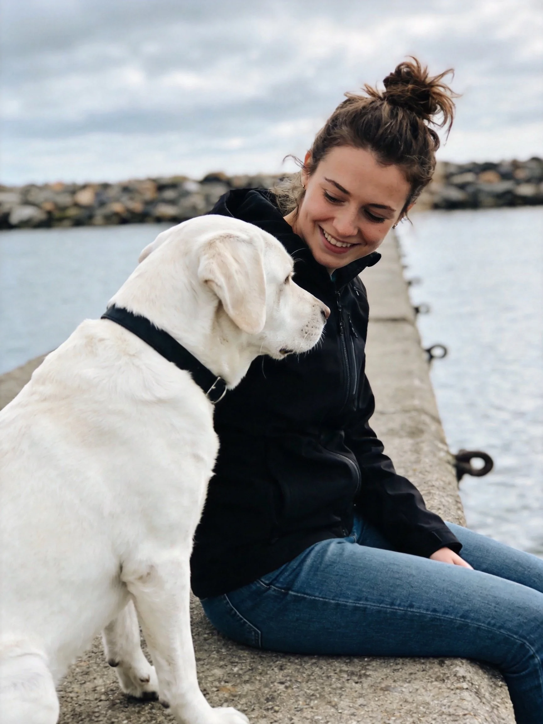 A woman in a black hoodie sits with a large white dog by the water.