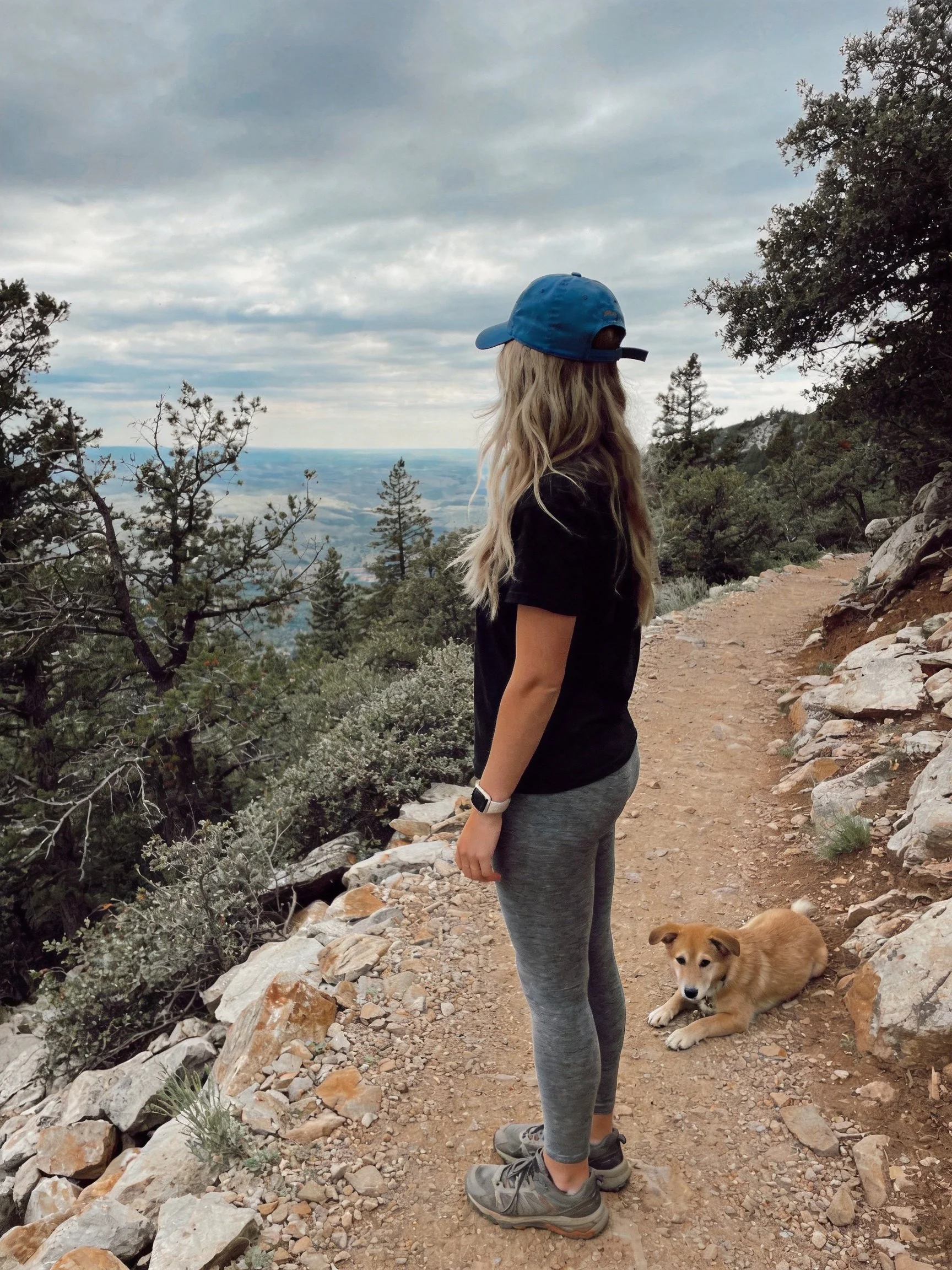 A woman in casual clothing stands beside a resting tan dog on a rocky hiking trail.