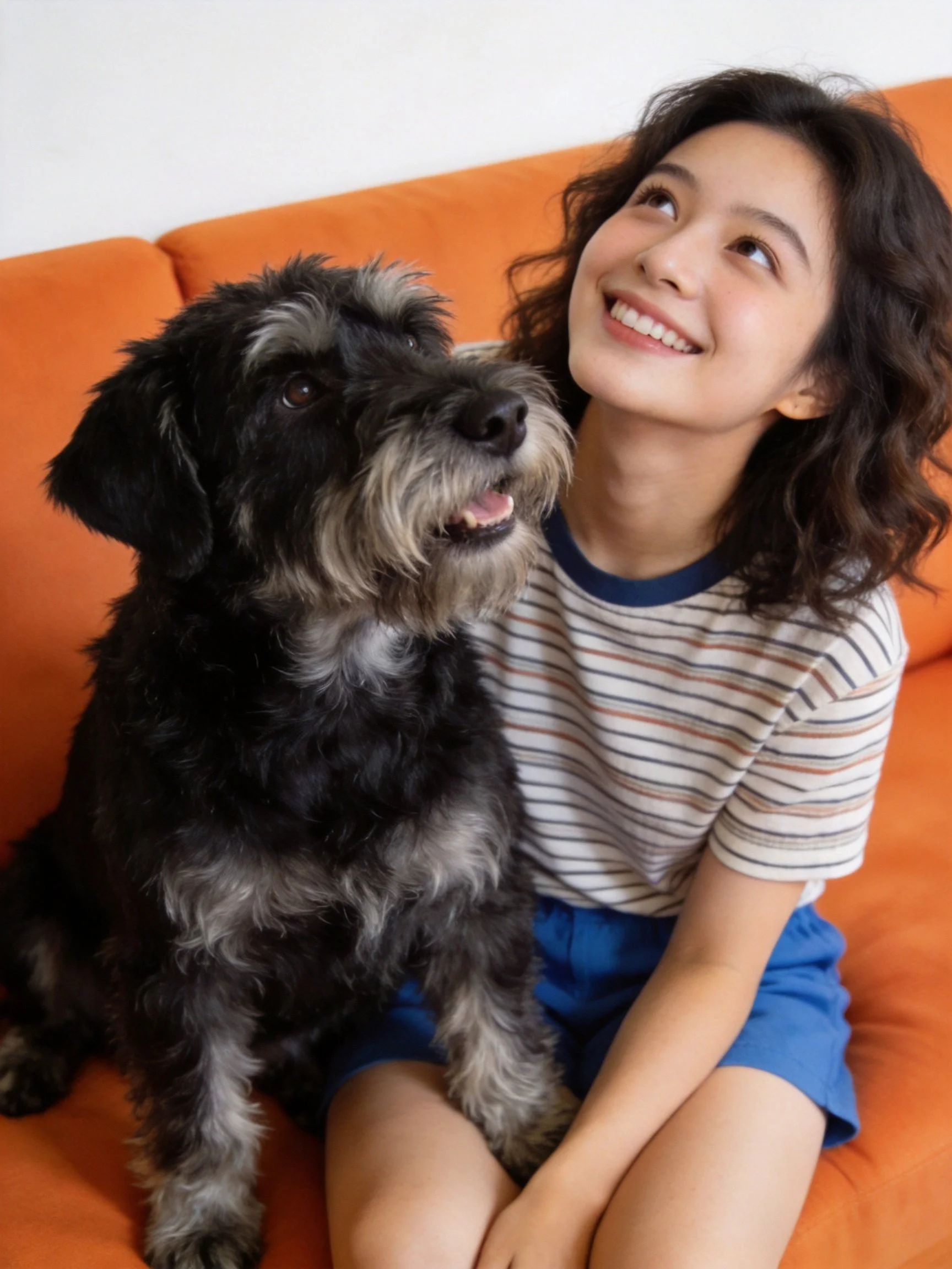 A woman sits on an orange couch with a black and gray dog, both looking upward.