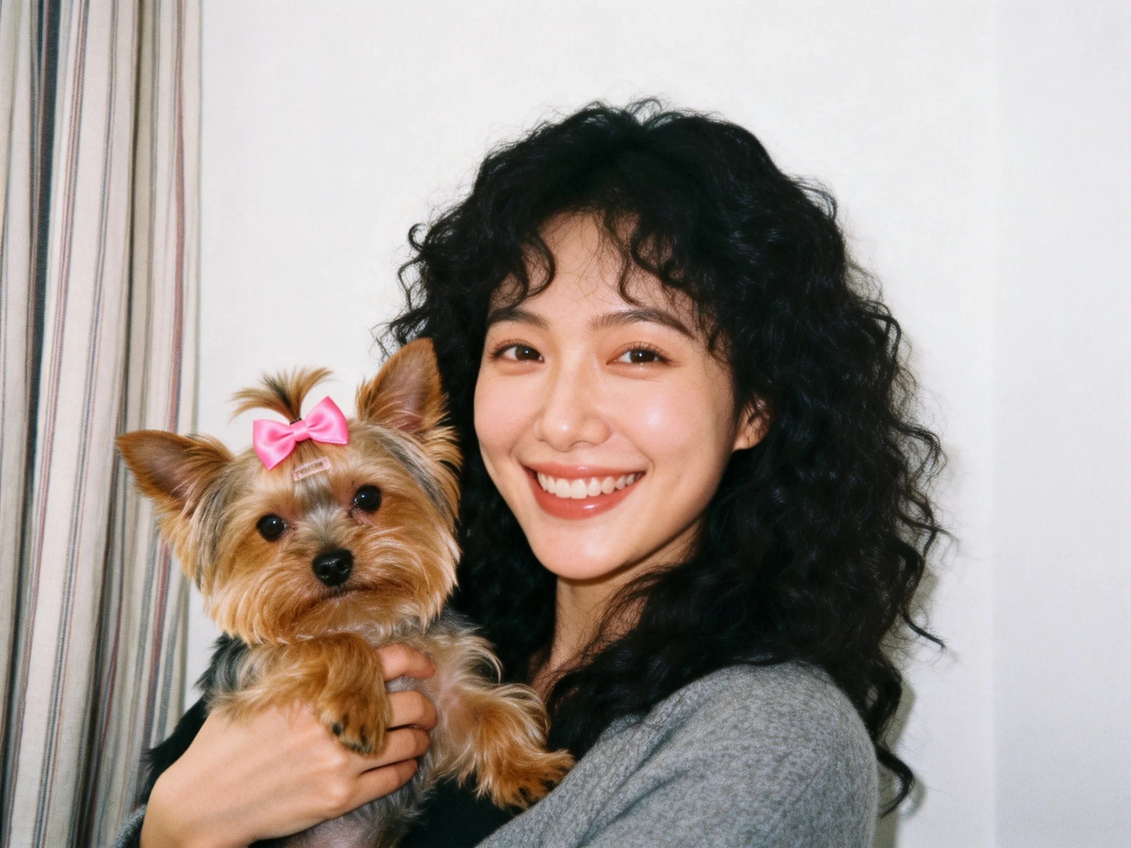 A woman smiles holding a small dog with a pink bow indoors