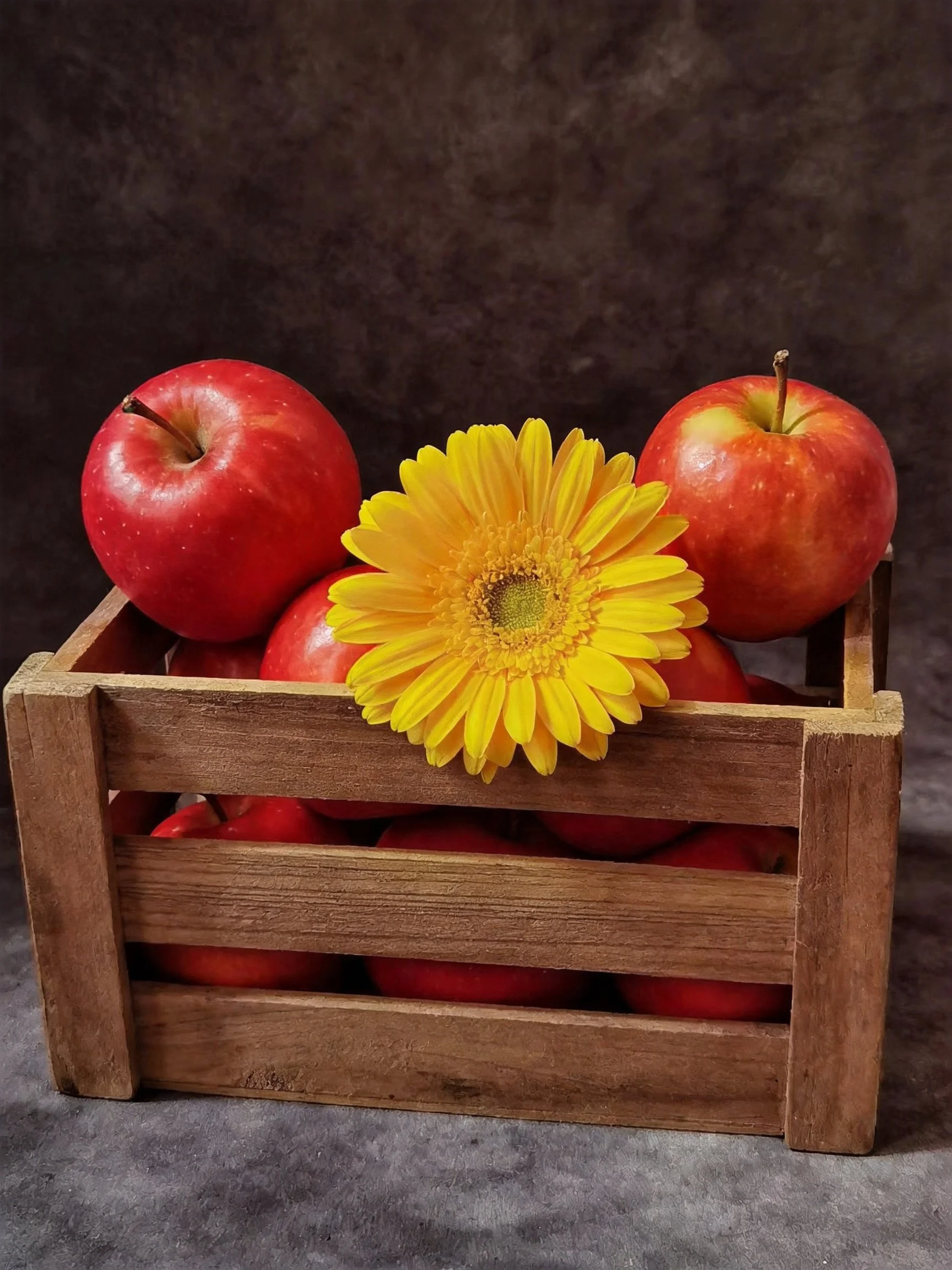 A wooden crate holds red apples and a vibrant yellow flower