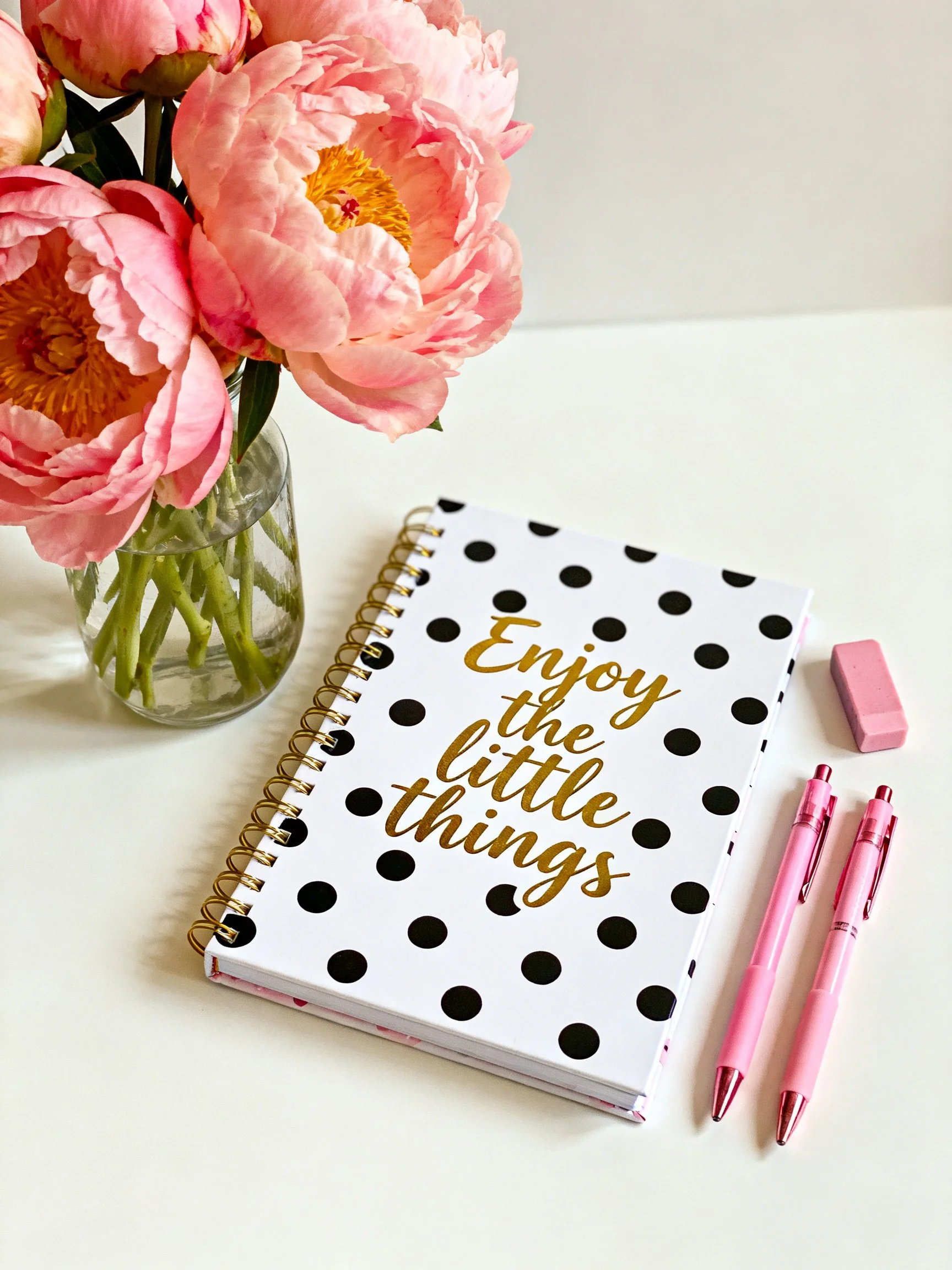 Pink peonies in glass vase beside polka dot notebook with inspirational text