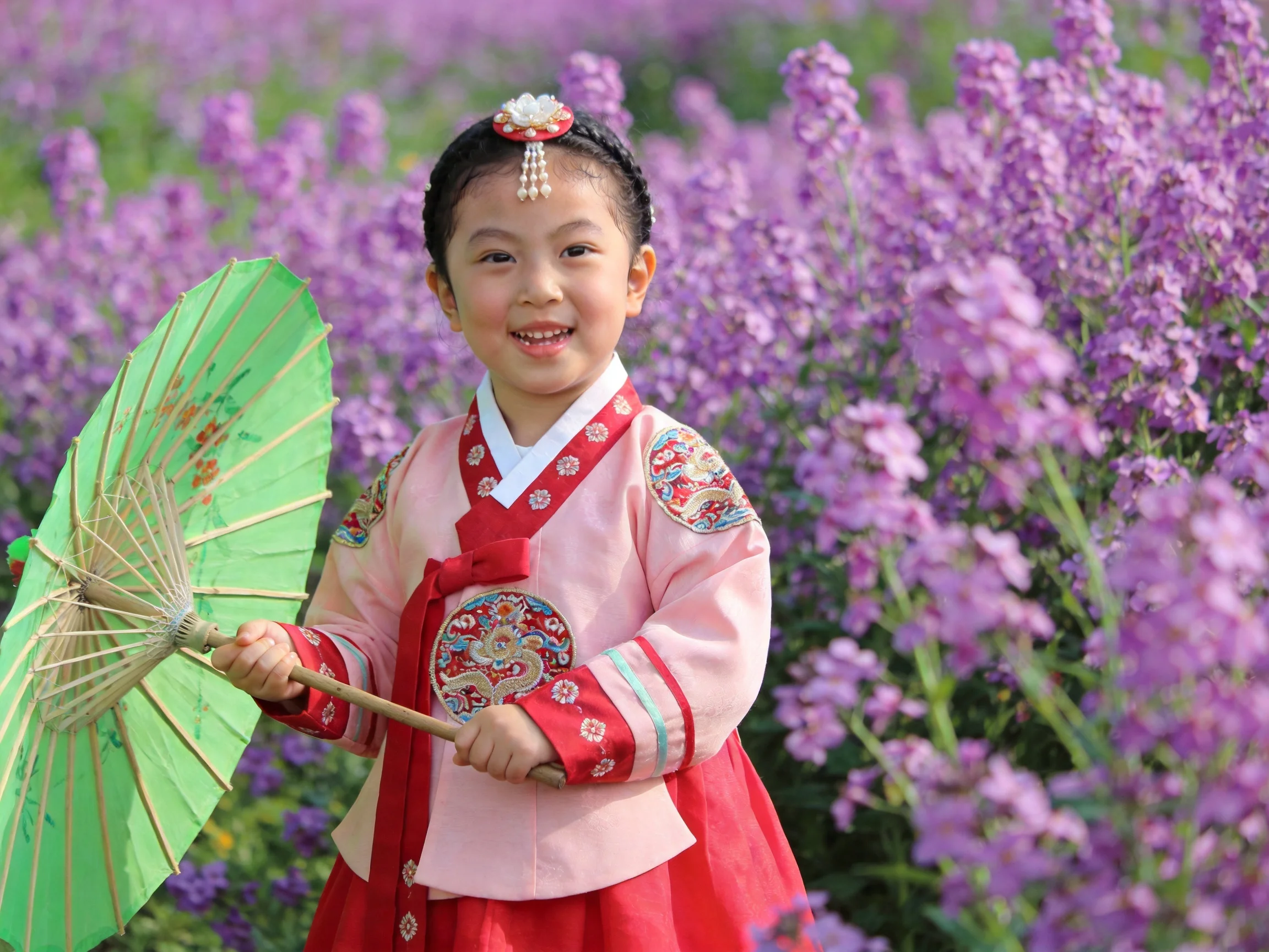 A child in traditional attire holds a green parasol among vibrant purple blooms