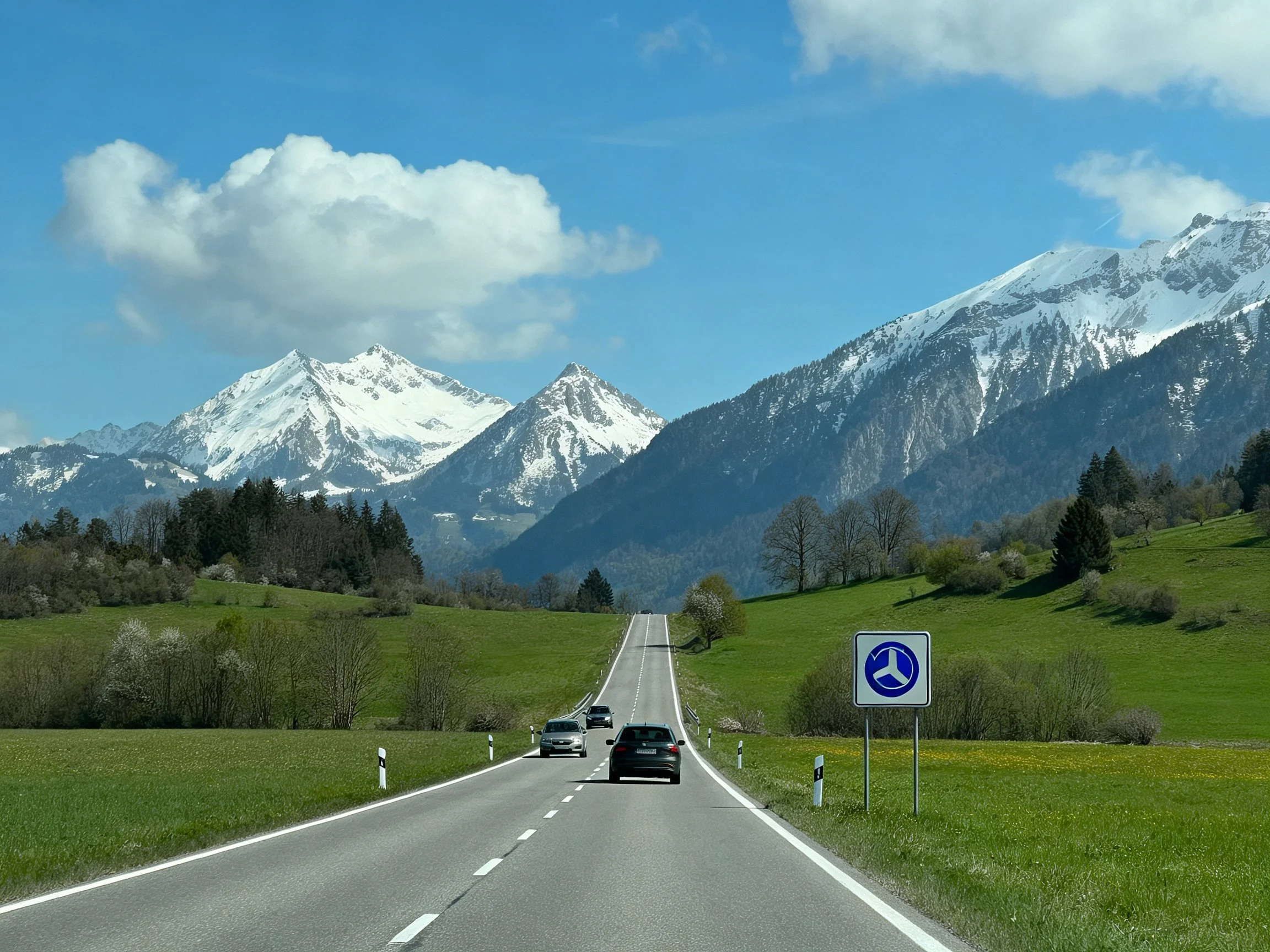 A highway winds through lush green fields toward snow-capped mountains