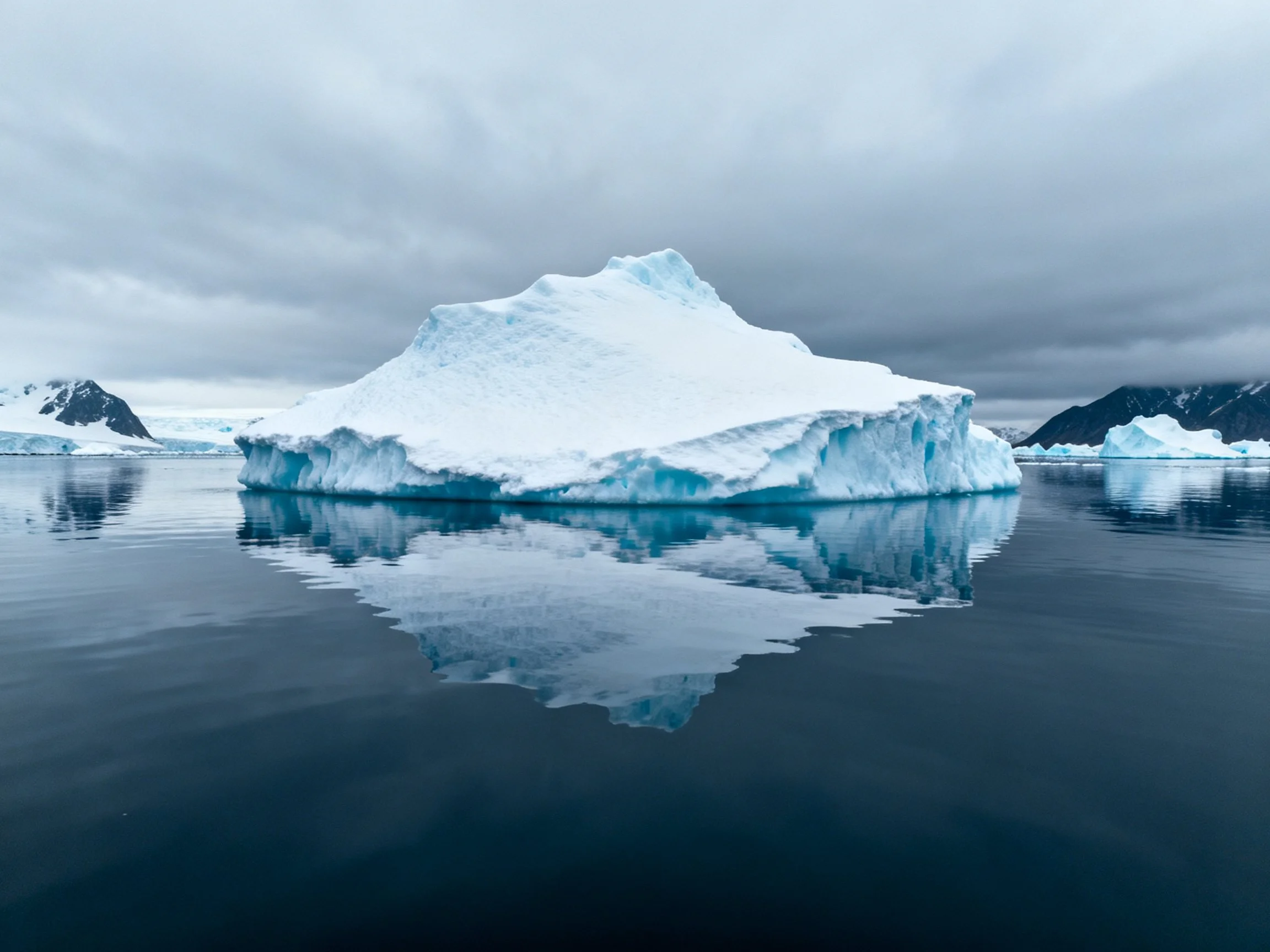 A large blue-tinted iceberg floats on calm water with mountain backdrop