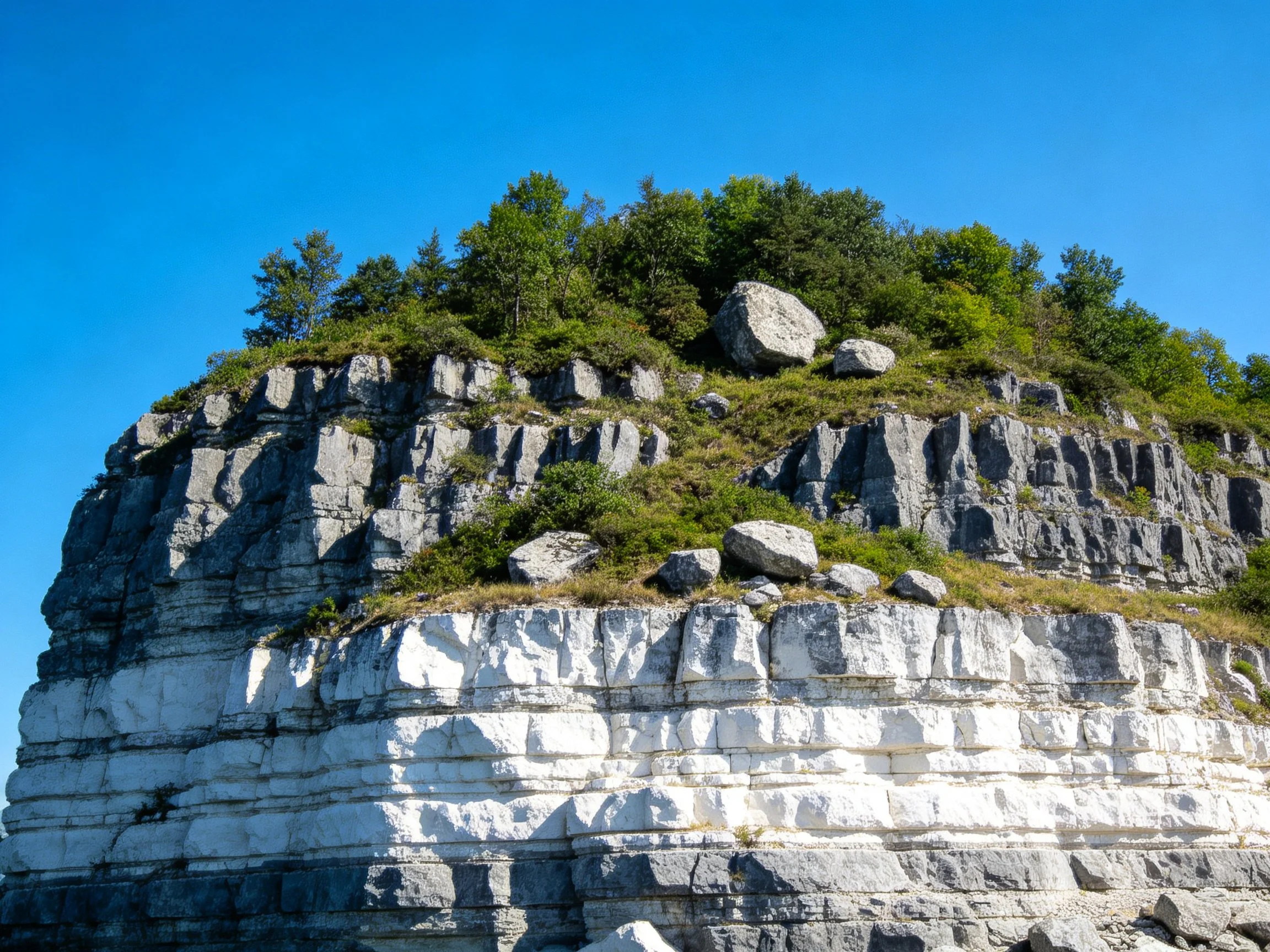 A layered limestone cliff with greenery and large boulders