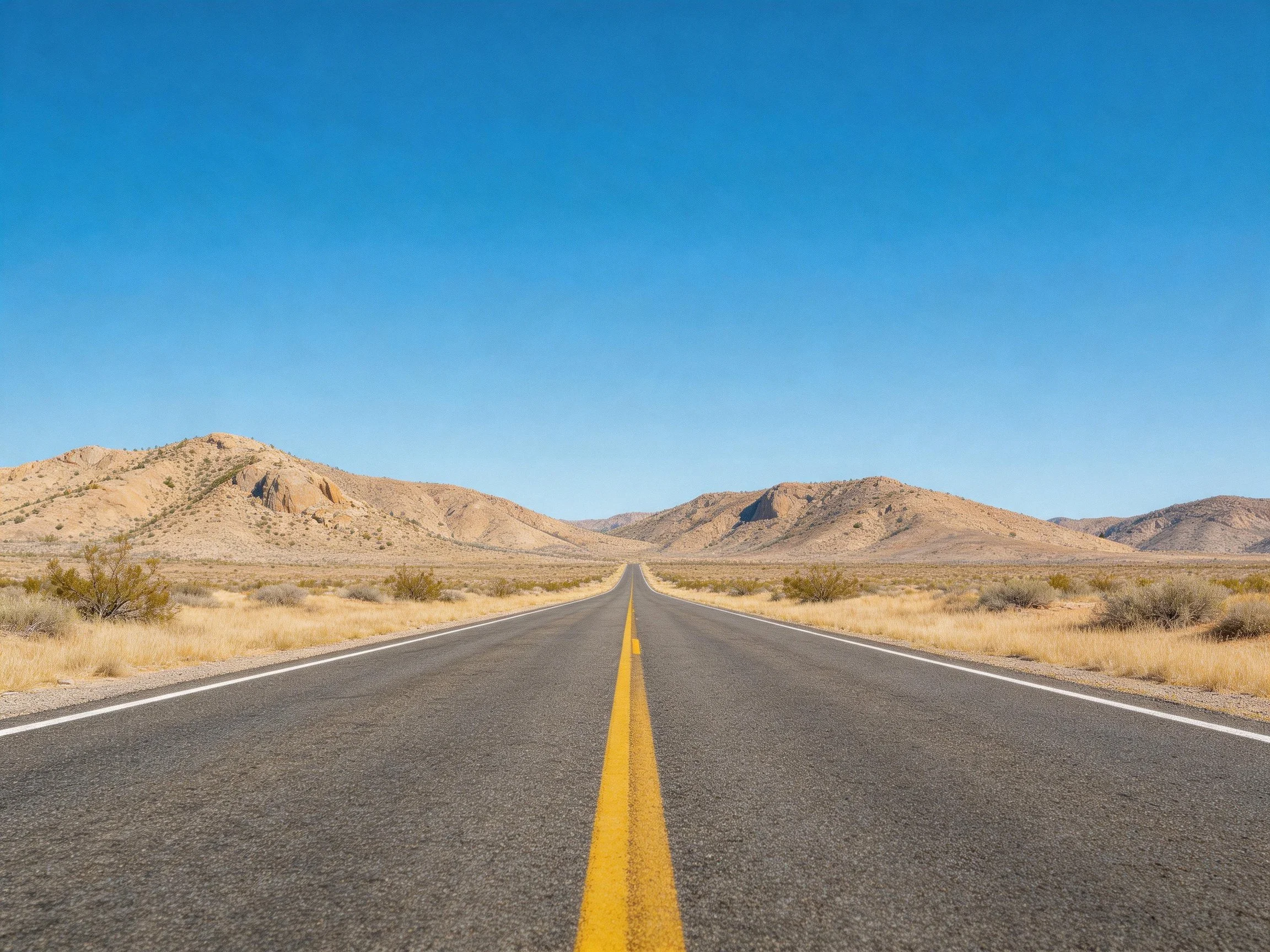 A long asphalt road leads toward rocky desert hills under clear blue sky