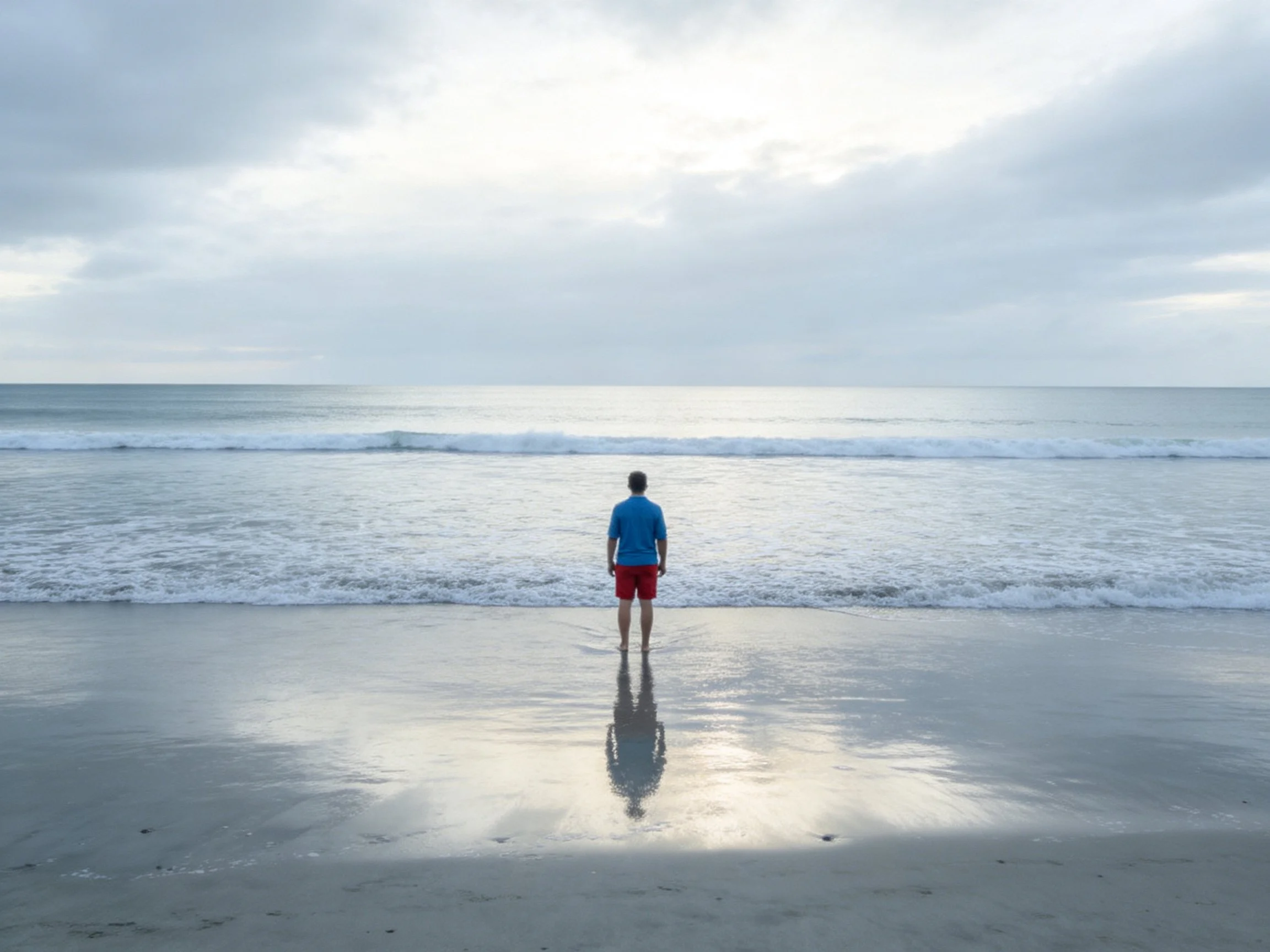 A man in blue shirt and red shorts stands at shoreline under cloudy sky
