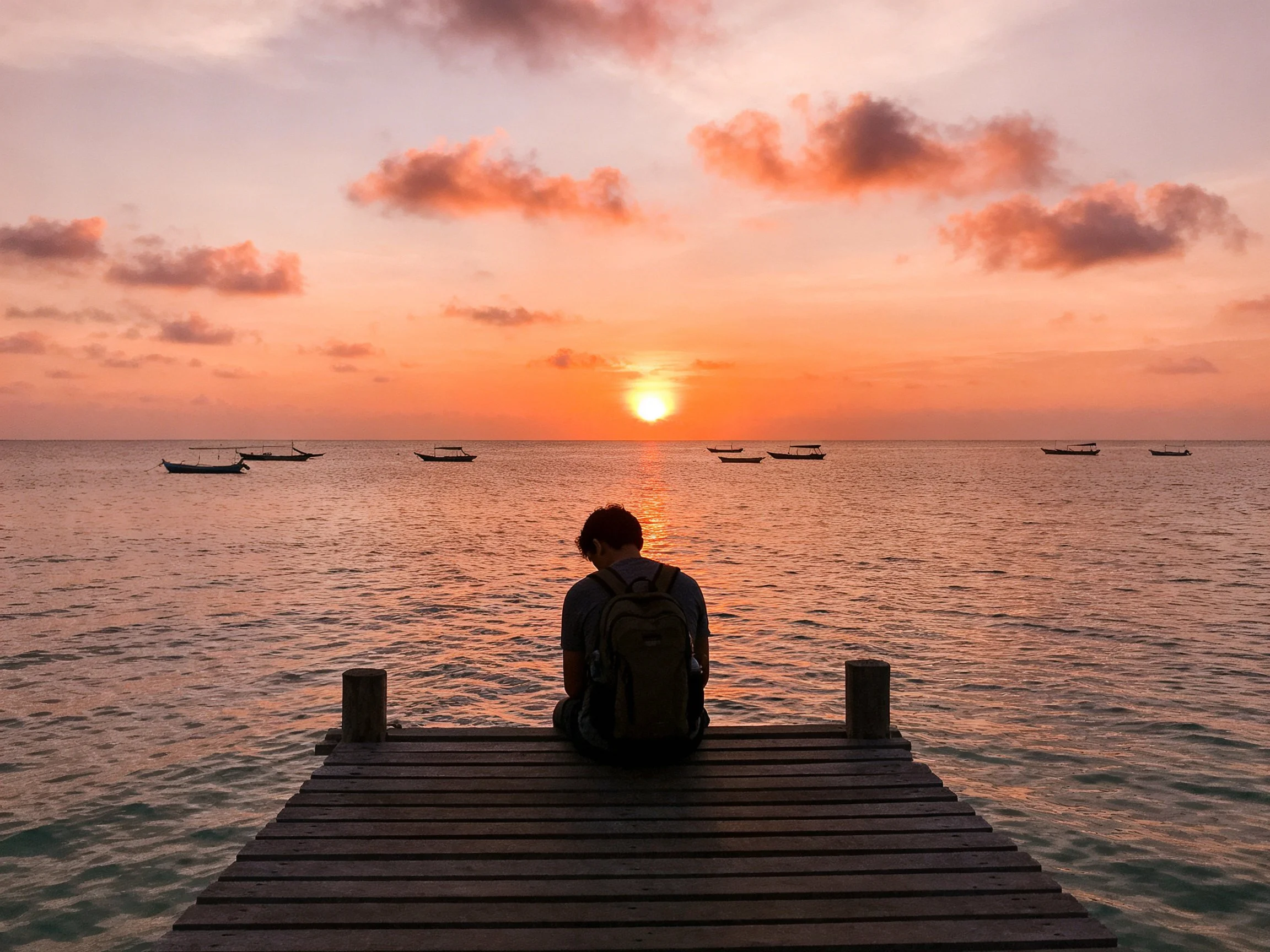 A person sits on a wooden dock viewing a vibrant orange sunset over calm waters
