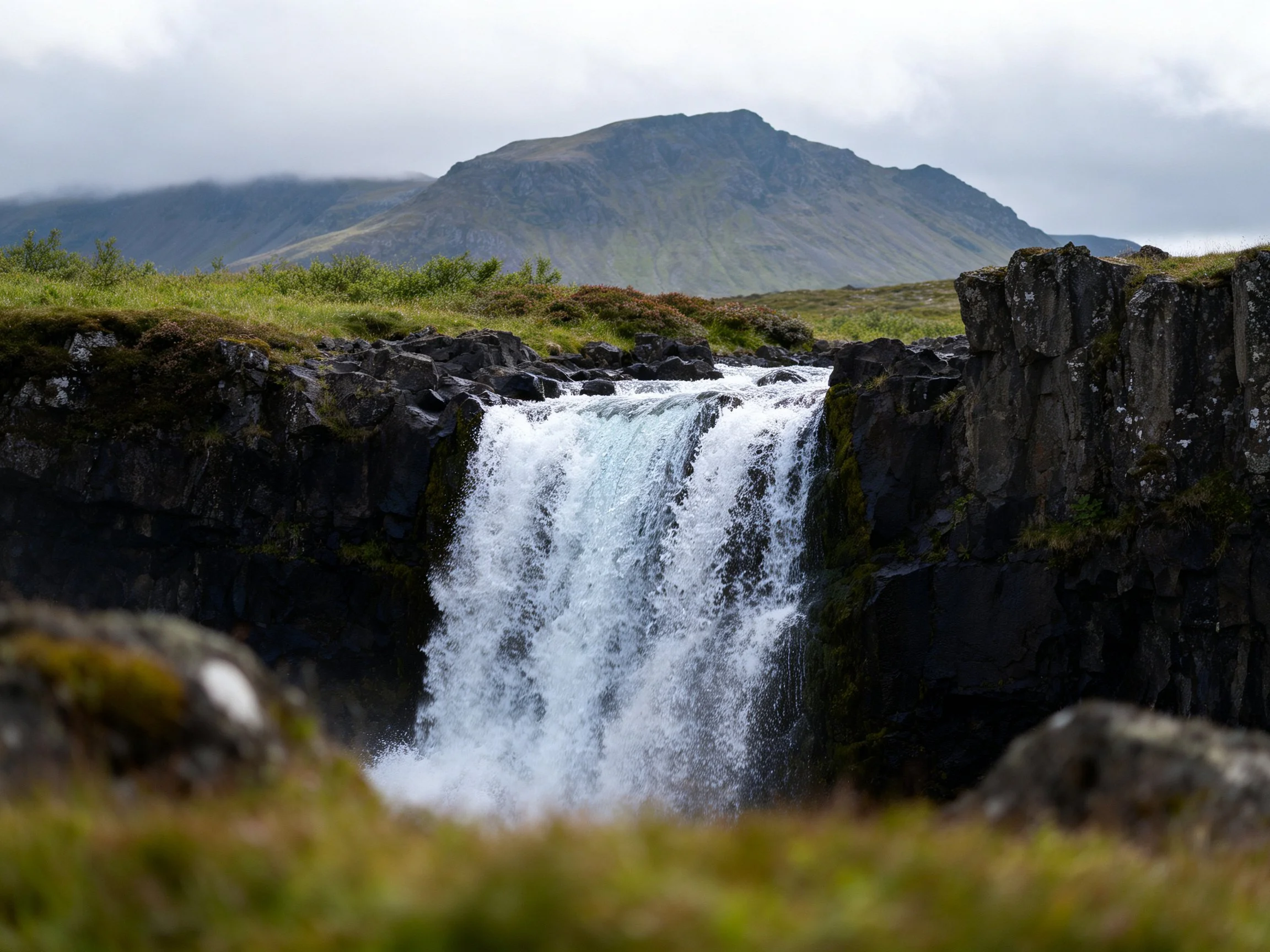 A powerful waterfall cascades between dark rocks near a misty mountain