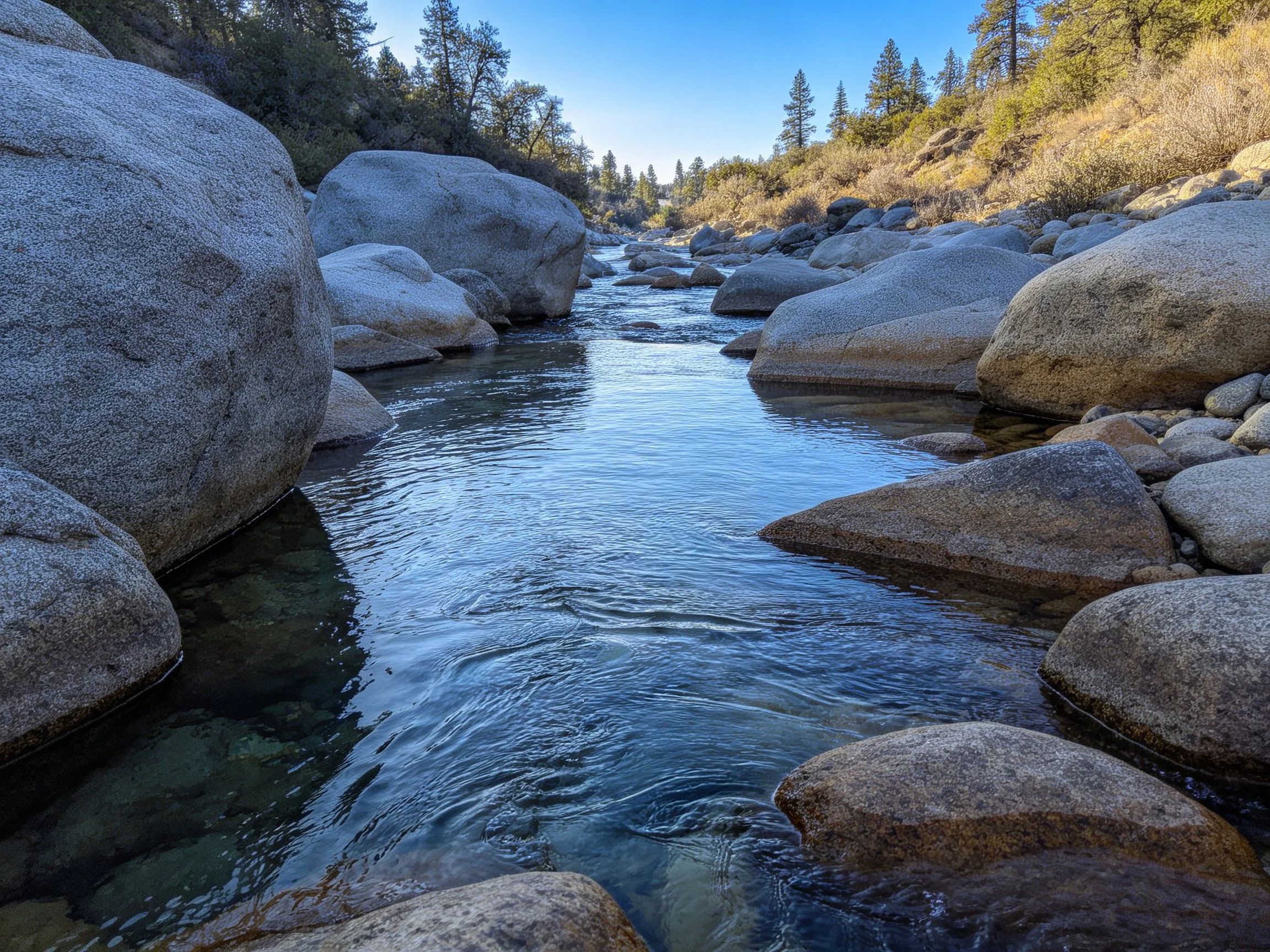 A rocky stream flows among large gray boulders near forested hills