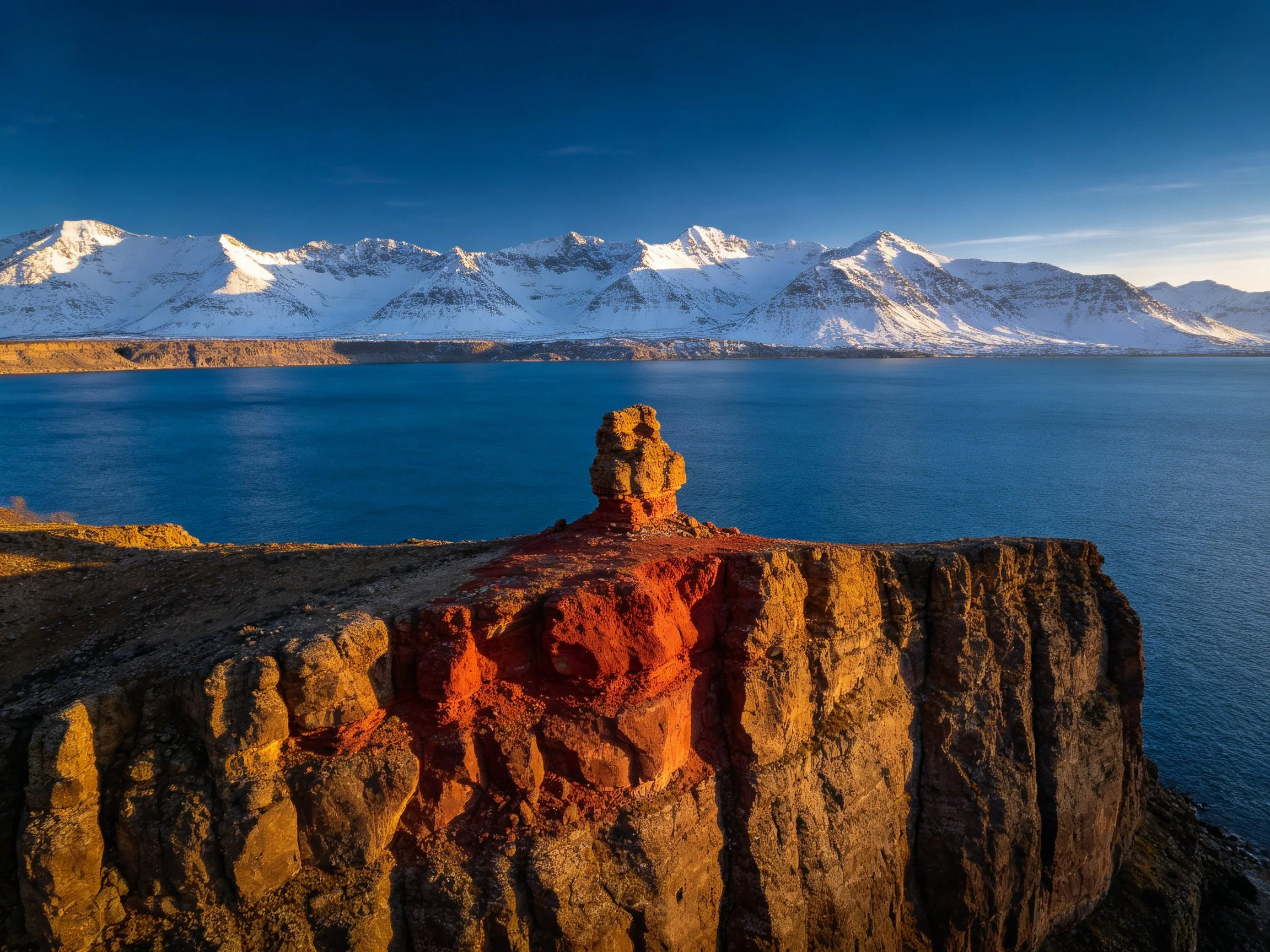 A rugged cliff with red rock formations overlooks a blue lake and snowy mountain range.