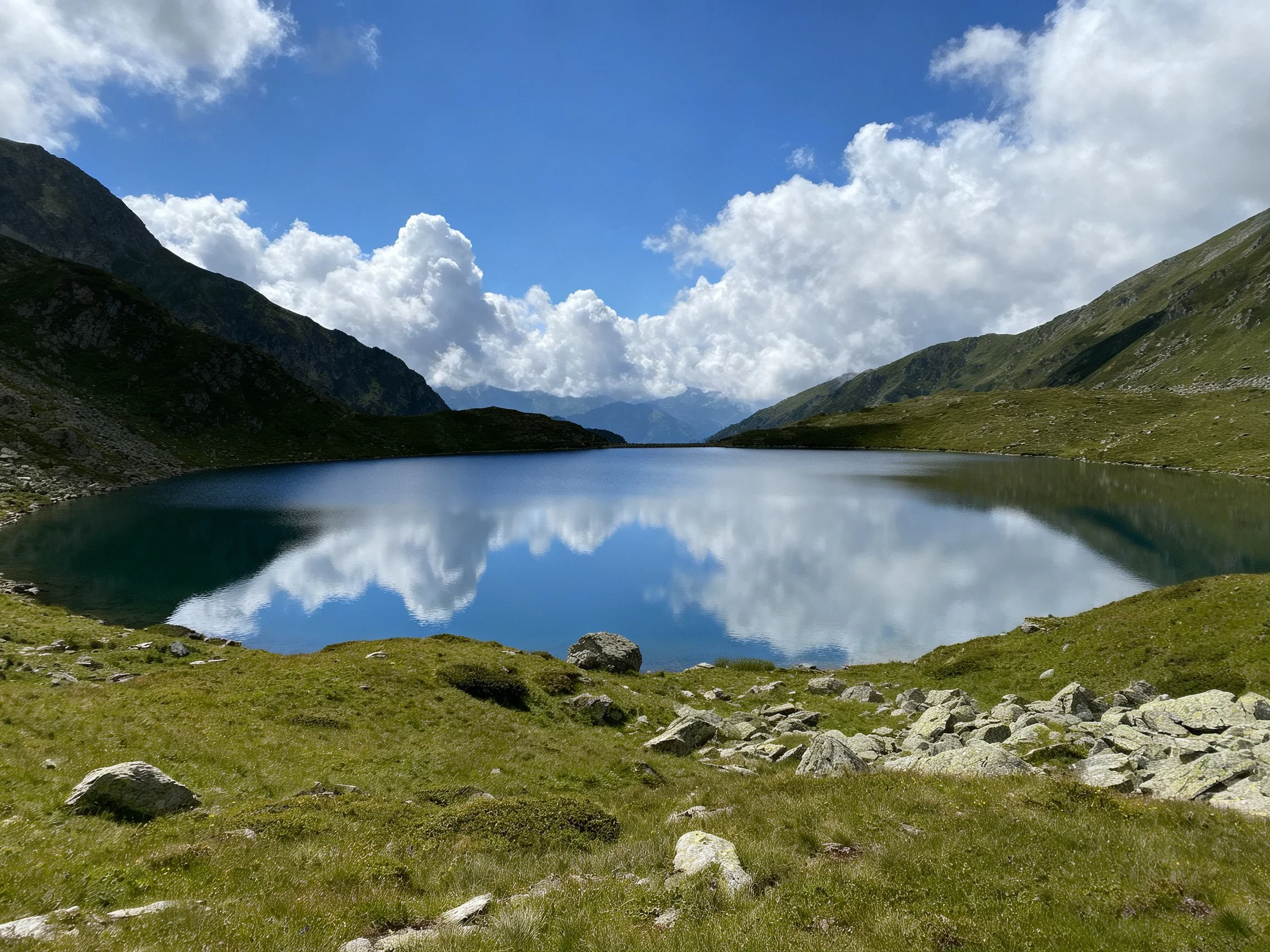 A serene alpine lake reflects blue sky and white clouds amid green mountains