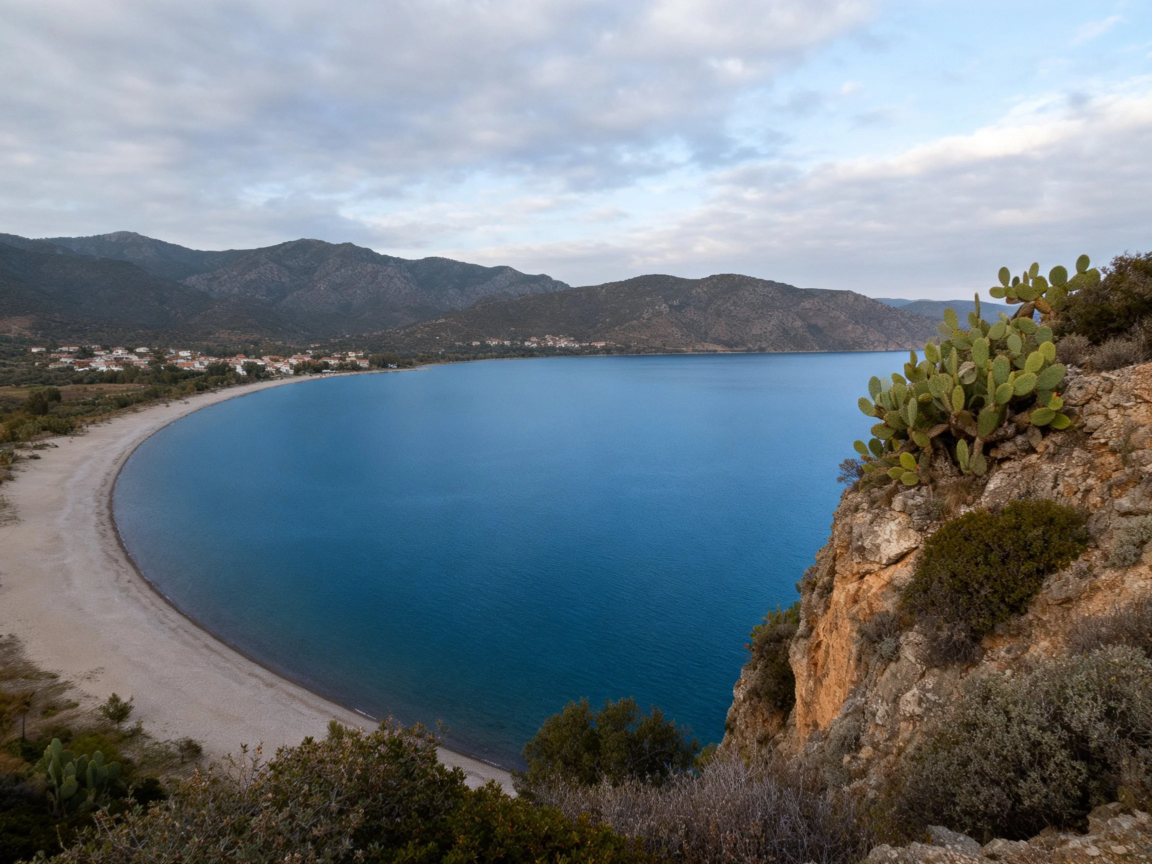 A serene bay with curved sandy shore, deep blue water, and mountainous backdrop.