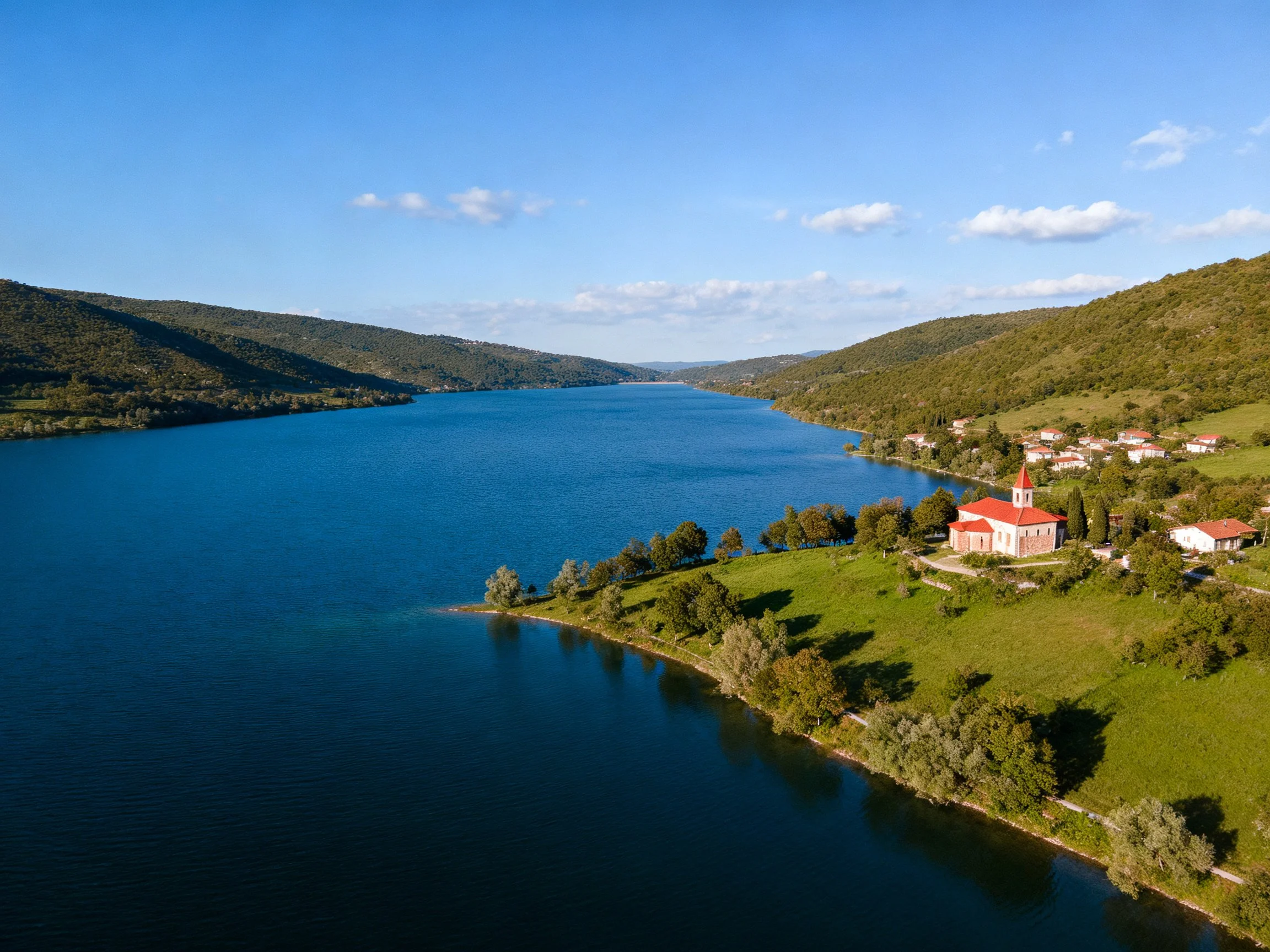 A serene blue lake bordered by rolling green hills and red-roofed settlements