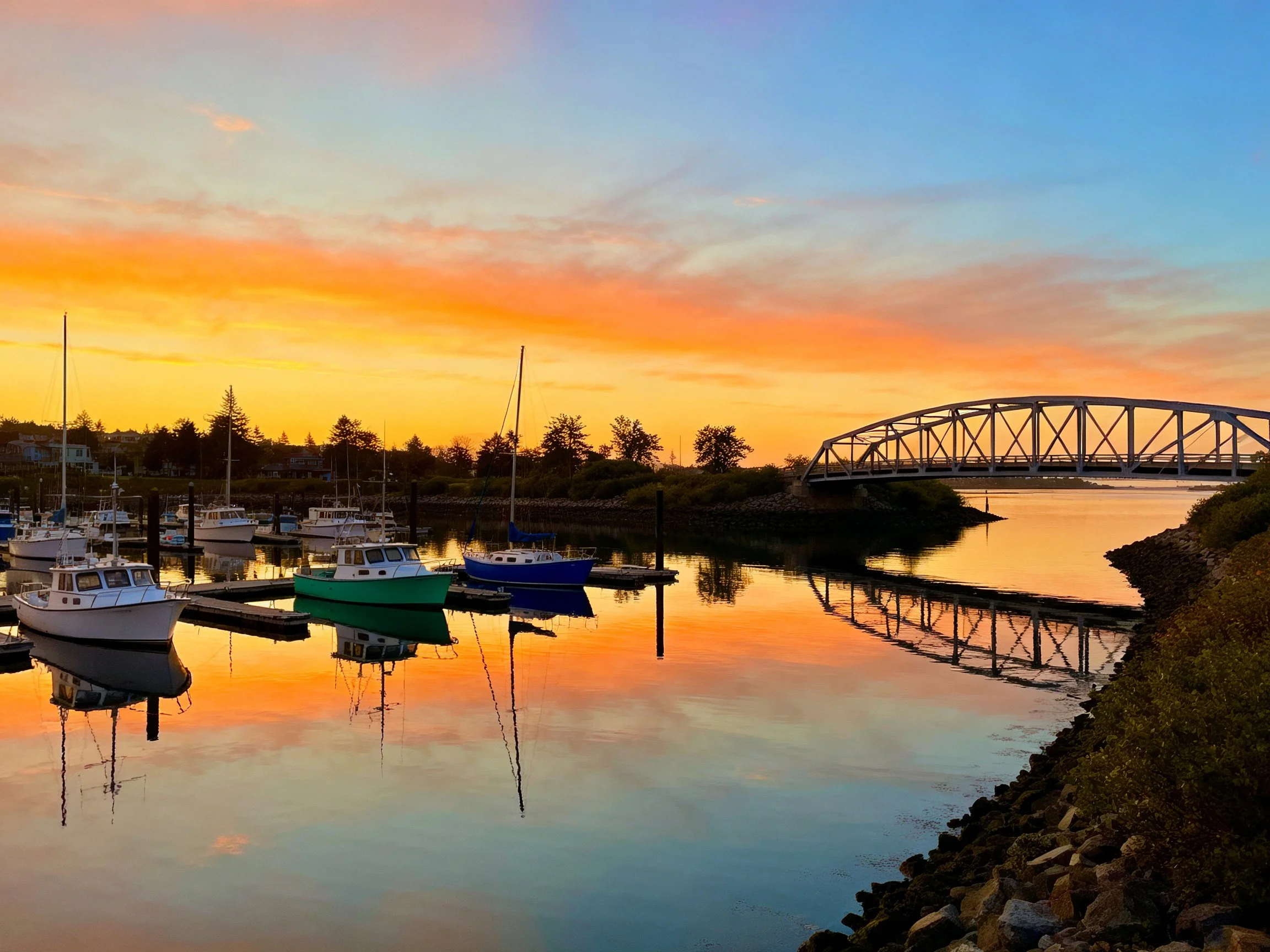 A serene harbor with colorful boats, a steel bridge, and vibrant sunset reflections