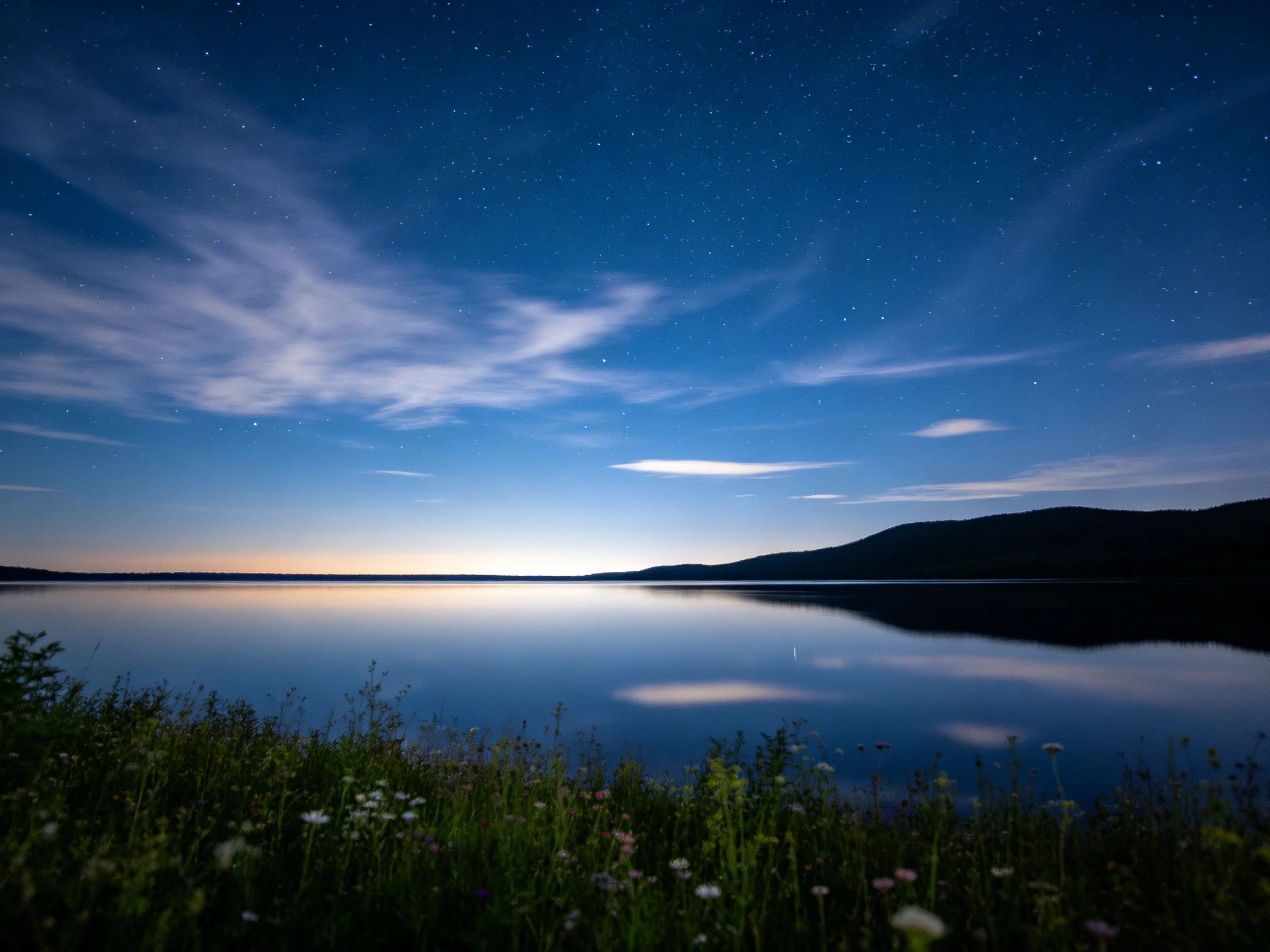 A starry twilight over a calm lake, distant mountains, and grassy shore