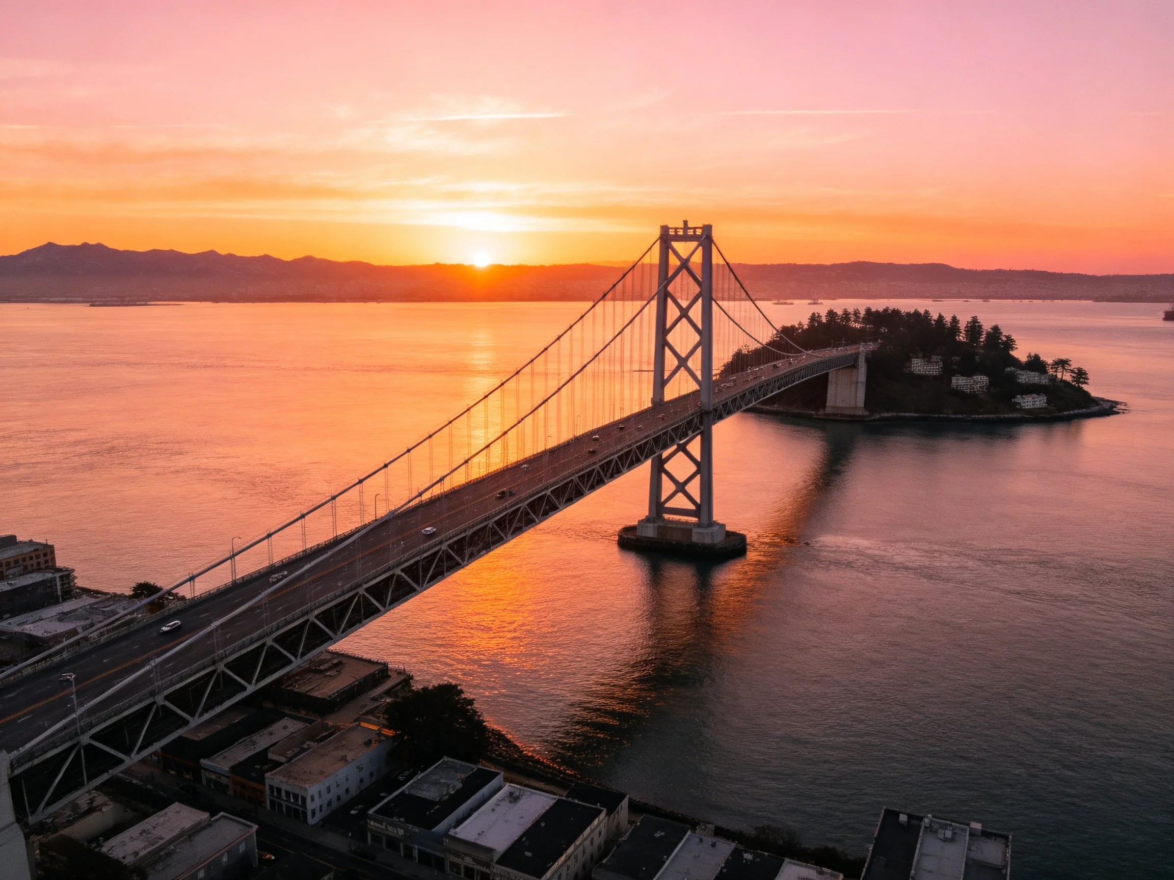 A tall suspension bridge spans calm bay waters at vibrant sunset