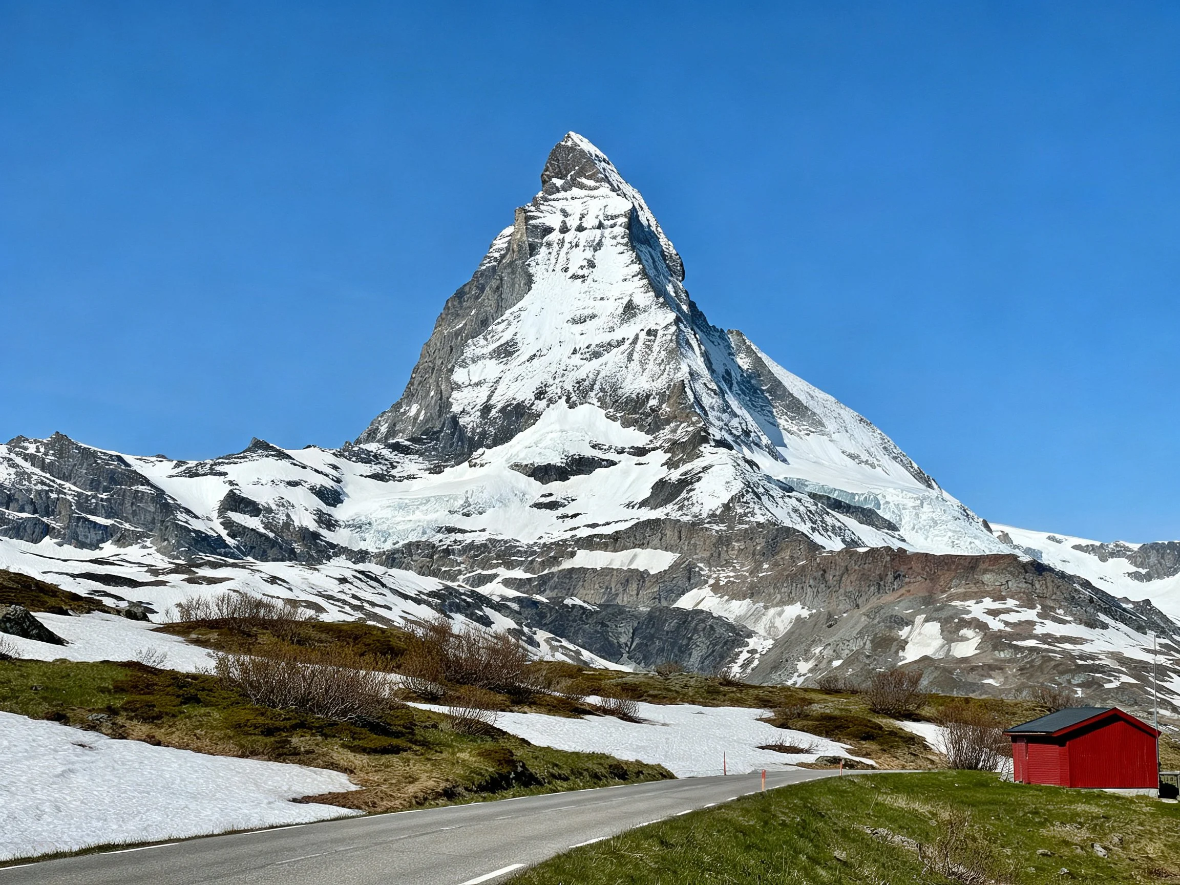 A towering snow - capped mountain with rocky slopes, green meadows, and red cabin