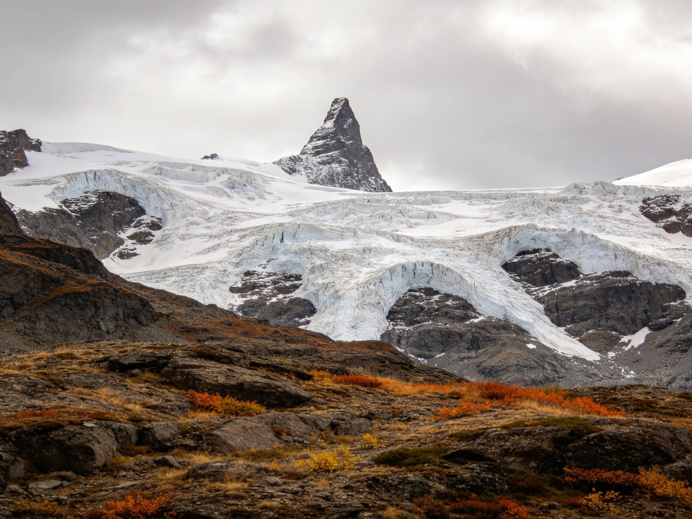 A triangular peak overlooks a sprawling glacier with rocky autumn terrain