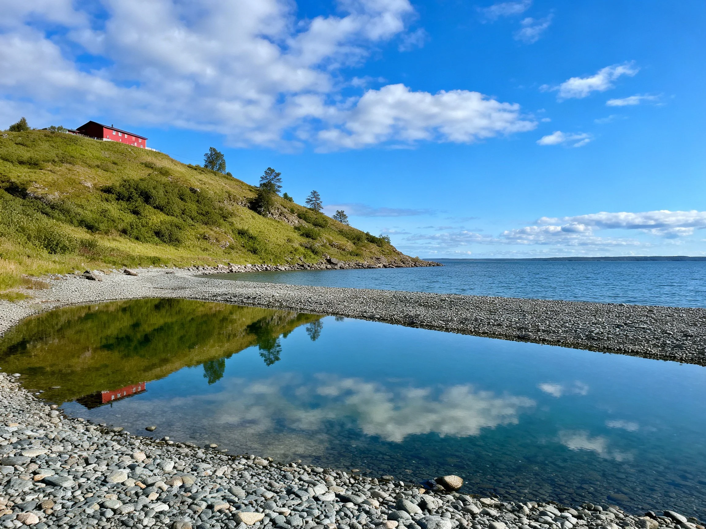 A vibrant red cabin overlooks a pebbly shoreline with reflective waters