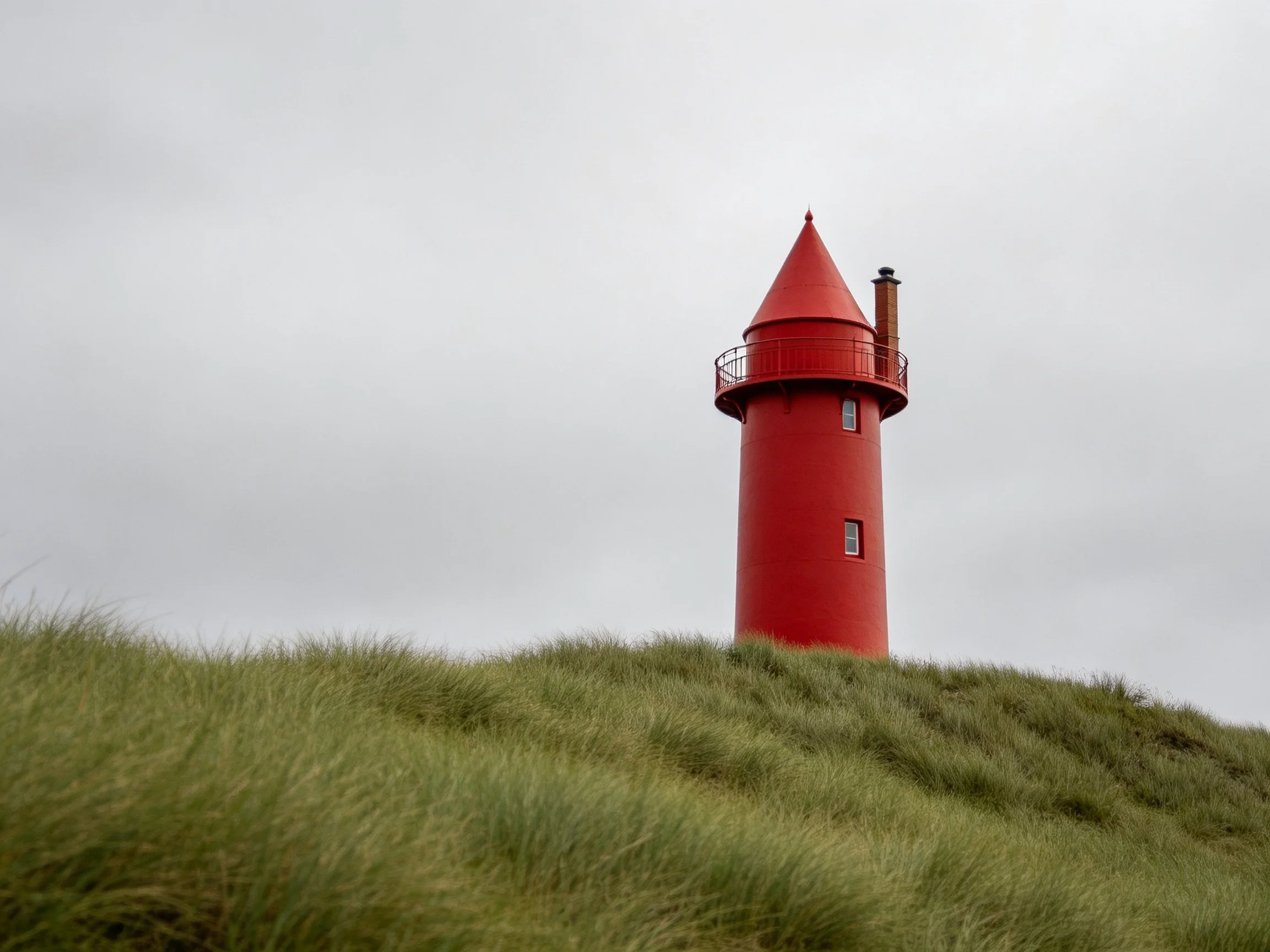 A vibrant red cylindrical tower atop a grassy hill under an overcast sky.