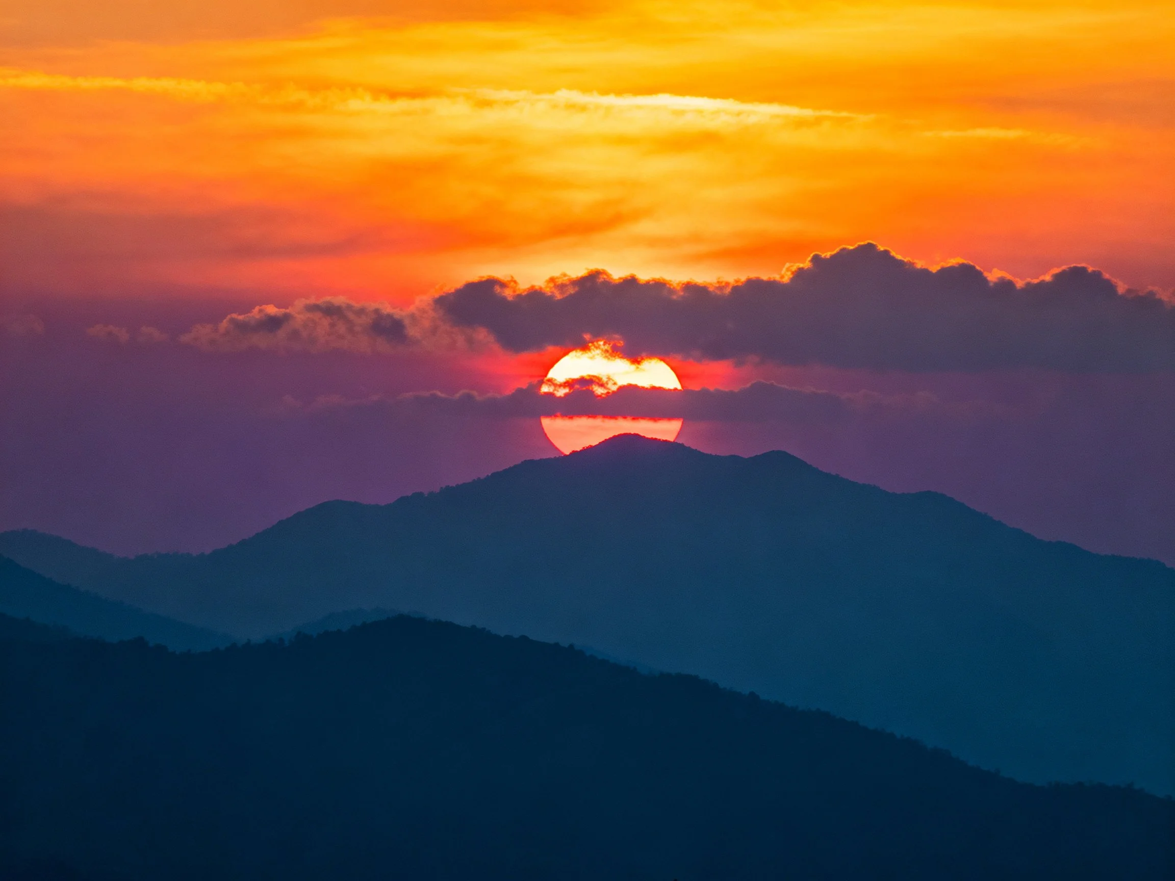 A vibrant sunset illuminates layered mountain silhouettes with orange-yellow-purple sky