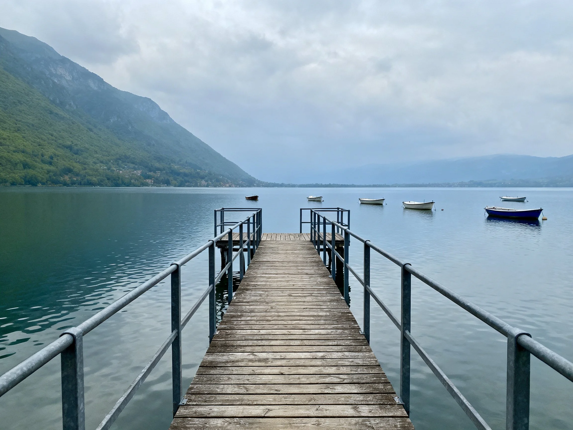A weathered wooden pier extends over calm lake with boats and mountains