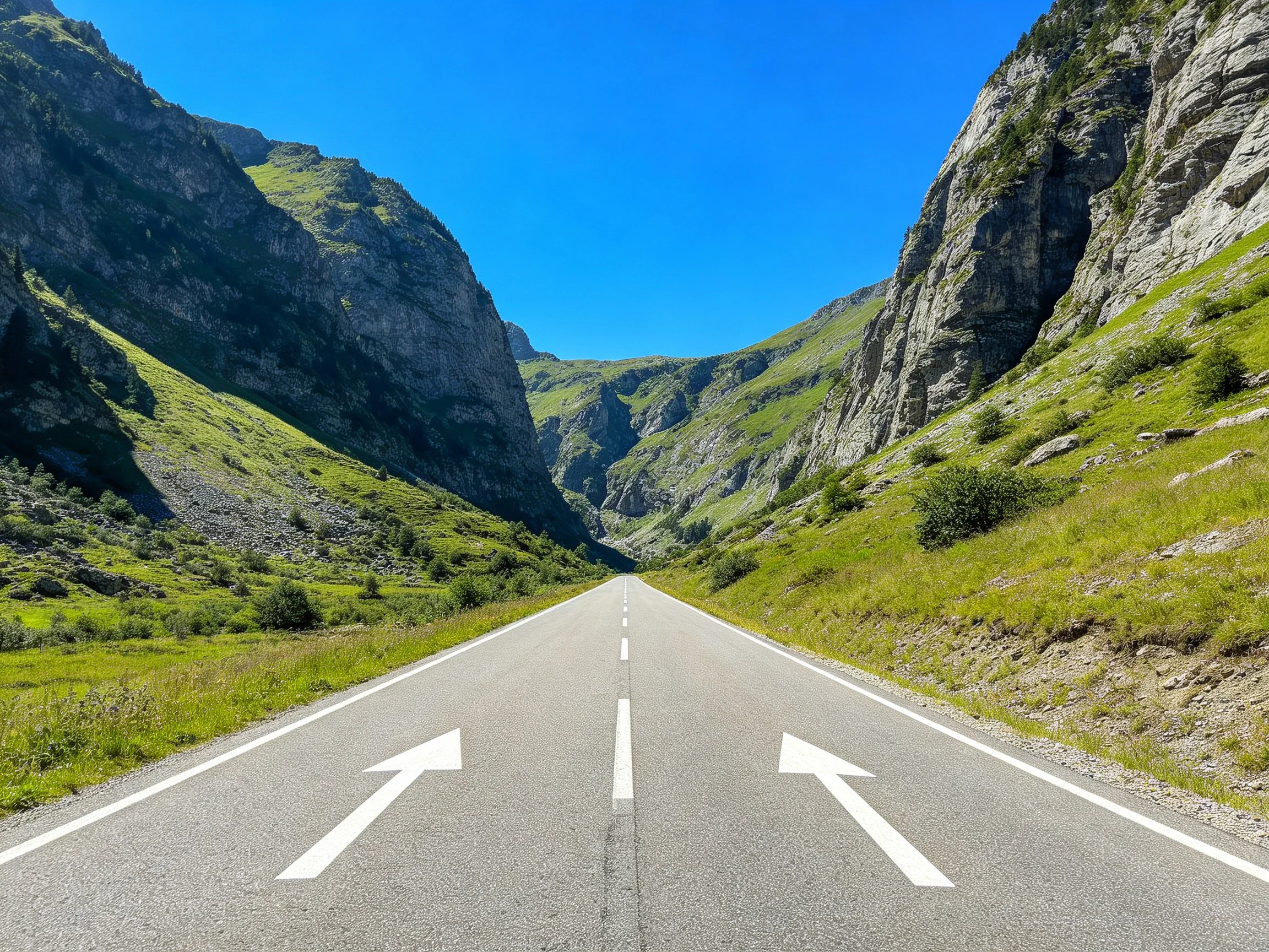A wide asphalt road flanked by steep rocky mountains with green slopes