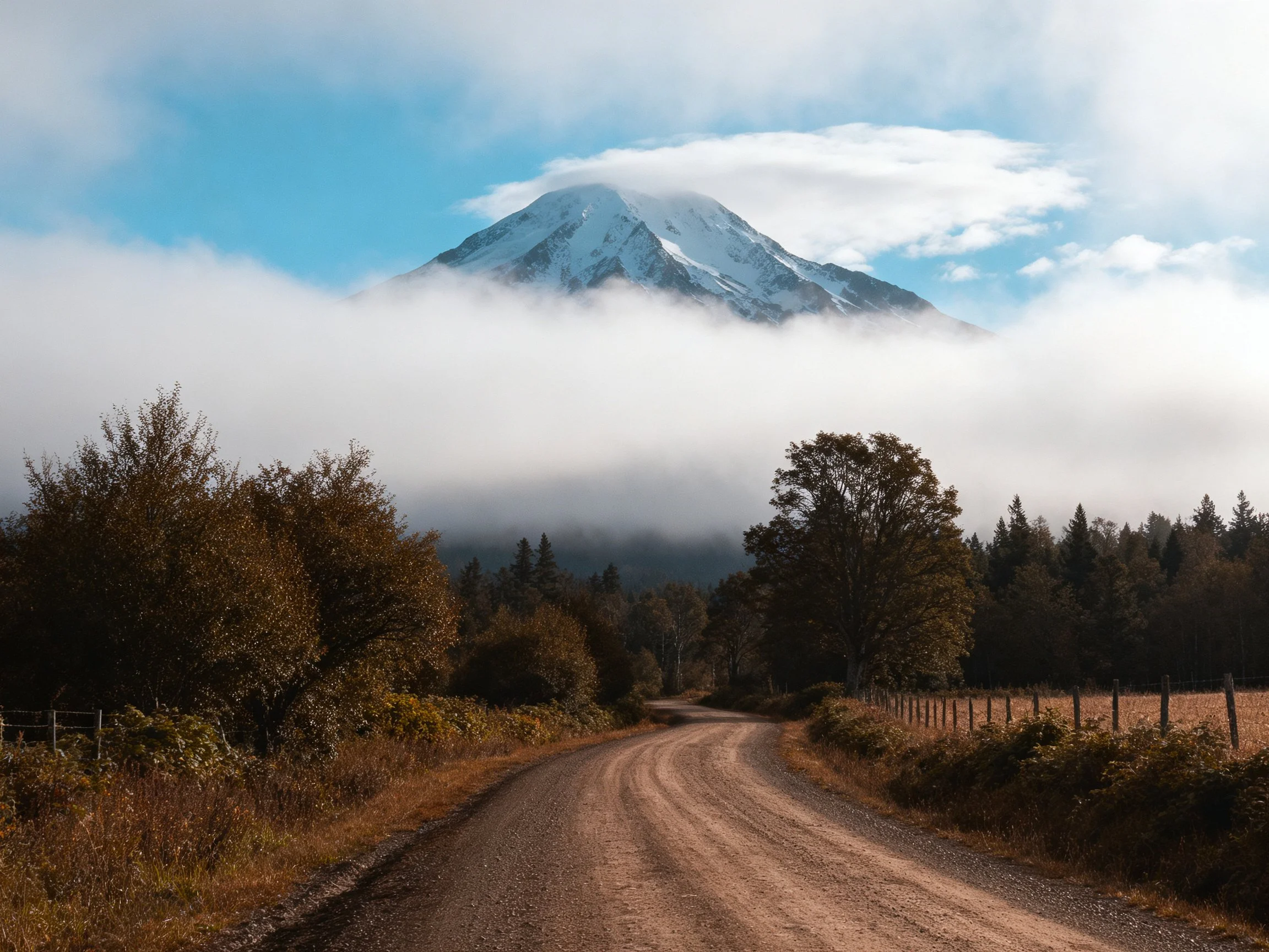 A winding dirt road leads toward a snow - capped mountain shrouded in fog