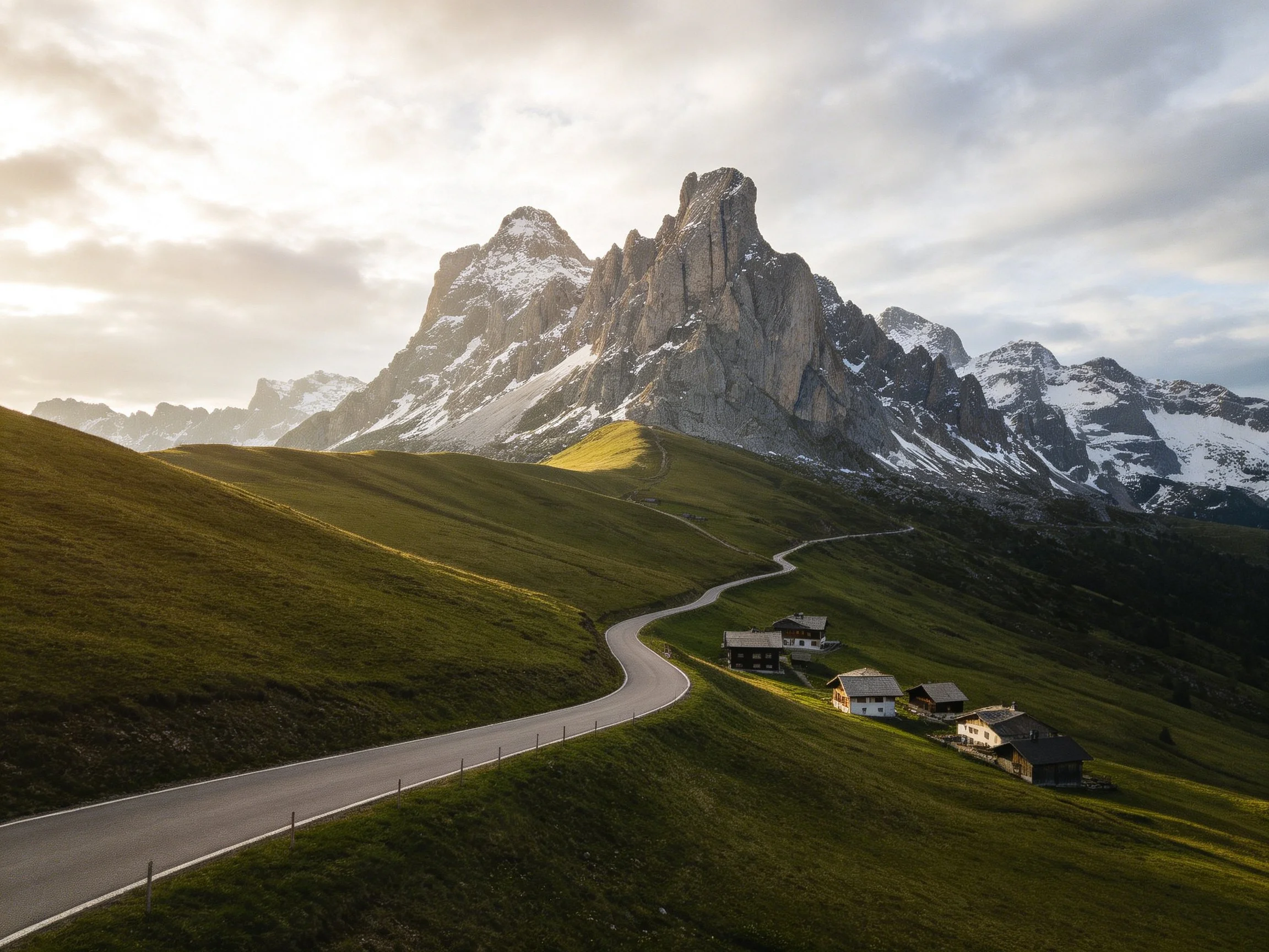A winding road leads past alpine houses near towering snow-capped peaks