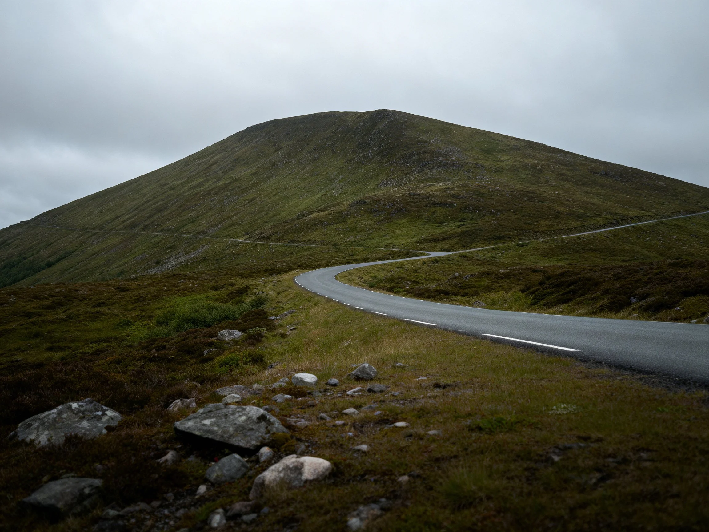 A winding road leads toward a large green hill under overcast skies