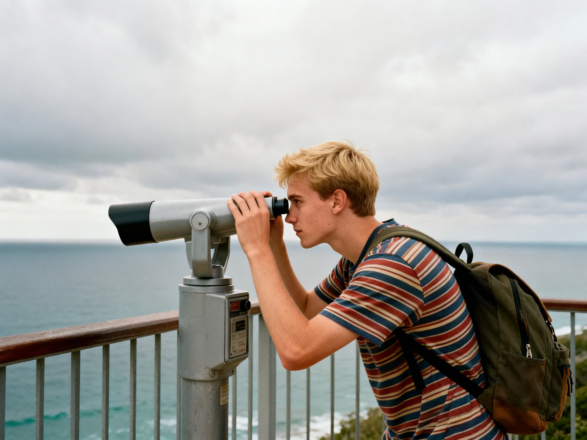 A young man with blonde hair uses binoculars at a coastal overlook under cloudy skies