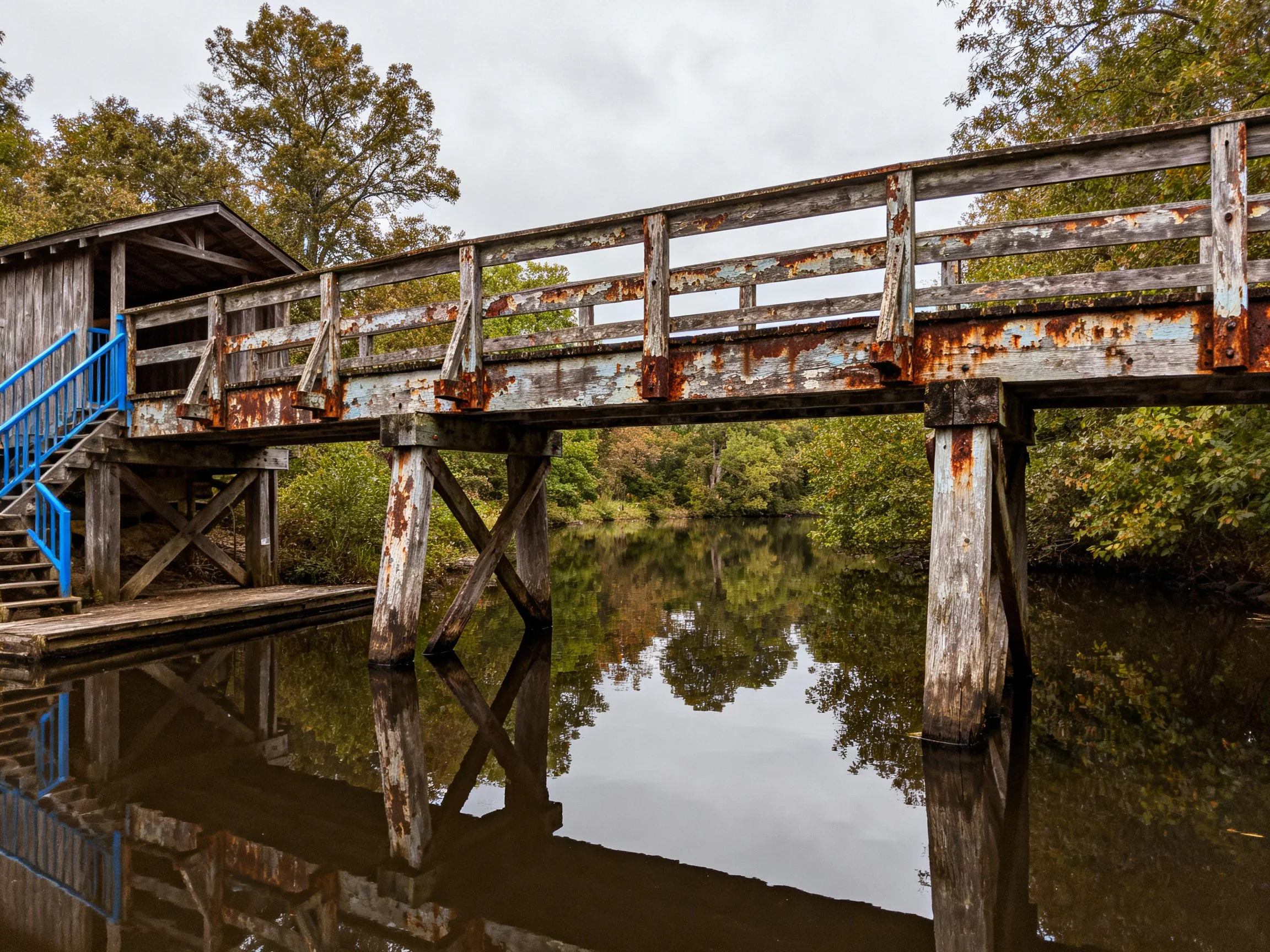 An old wooden bridge with rusted railings spans still water near blue stairs