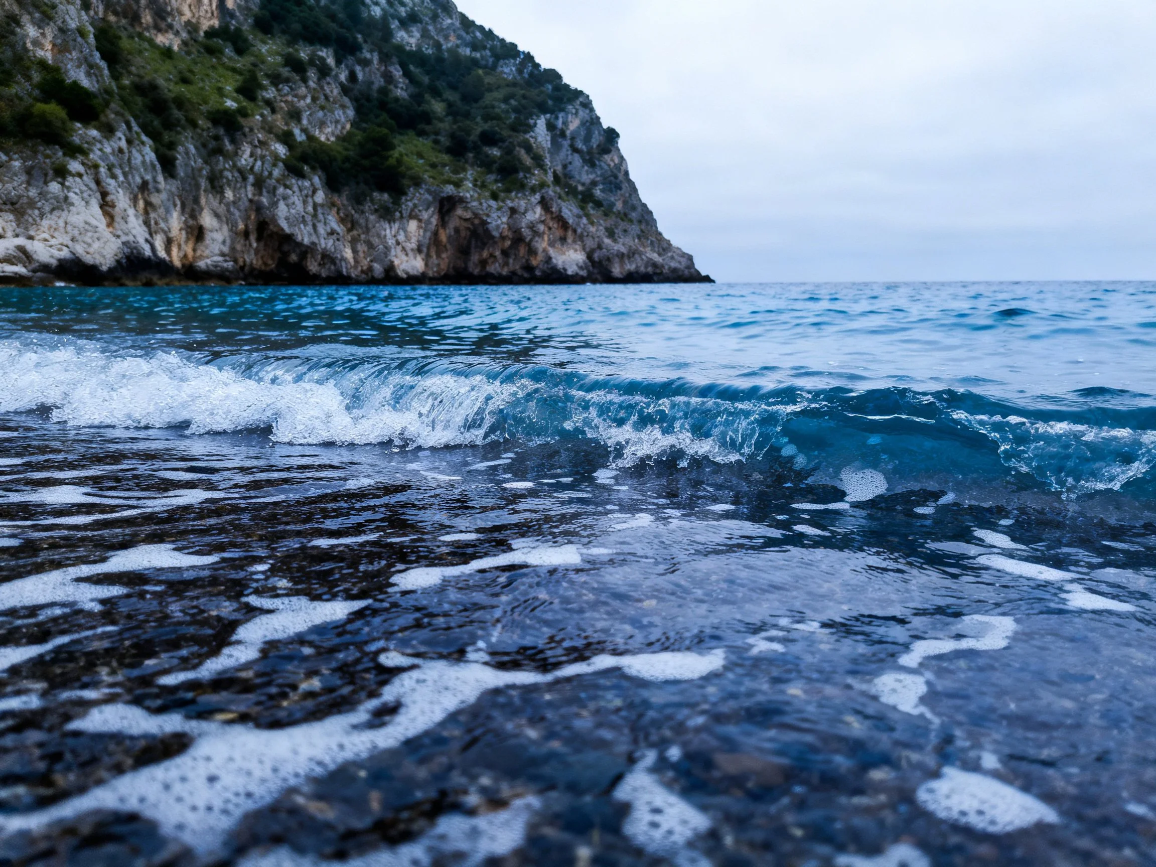 Blue Ocean Waves Strike Rocky Beach Near Green-Covered Cliffs